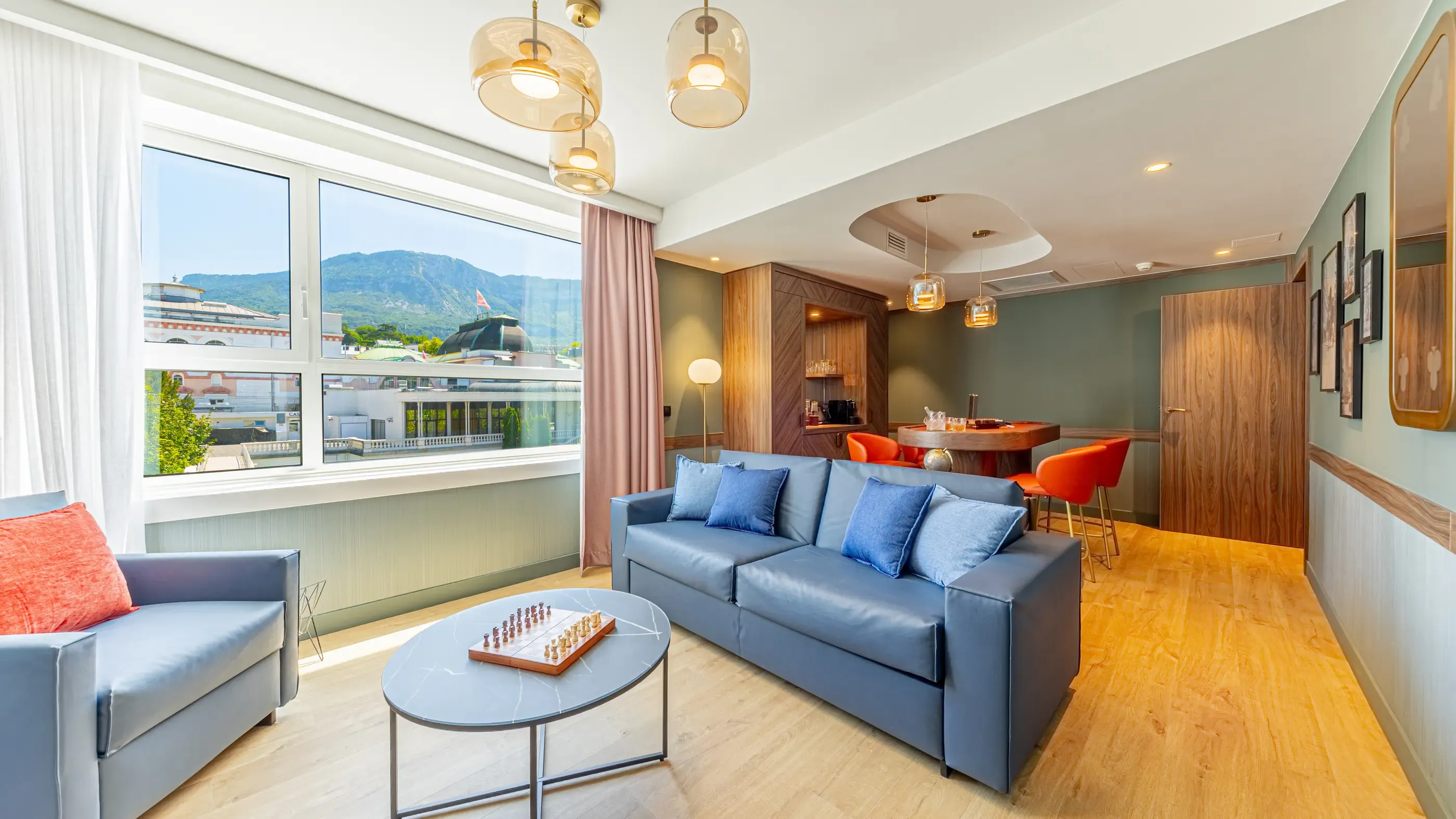 Salon moderne avec canapé bleu, table basse, échiquier et vue panoramique sur une ville et des montagnes