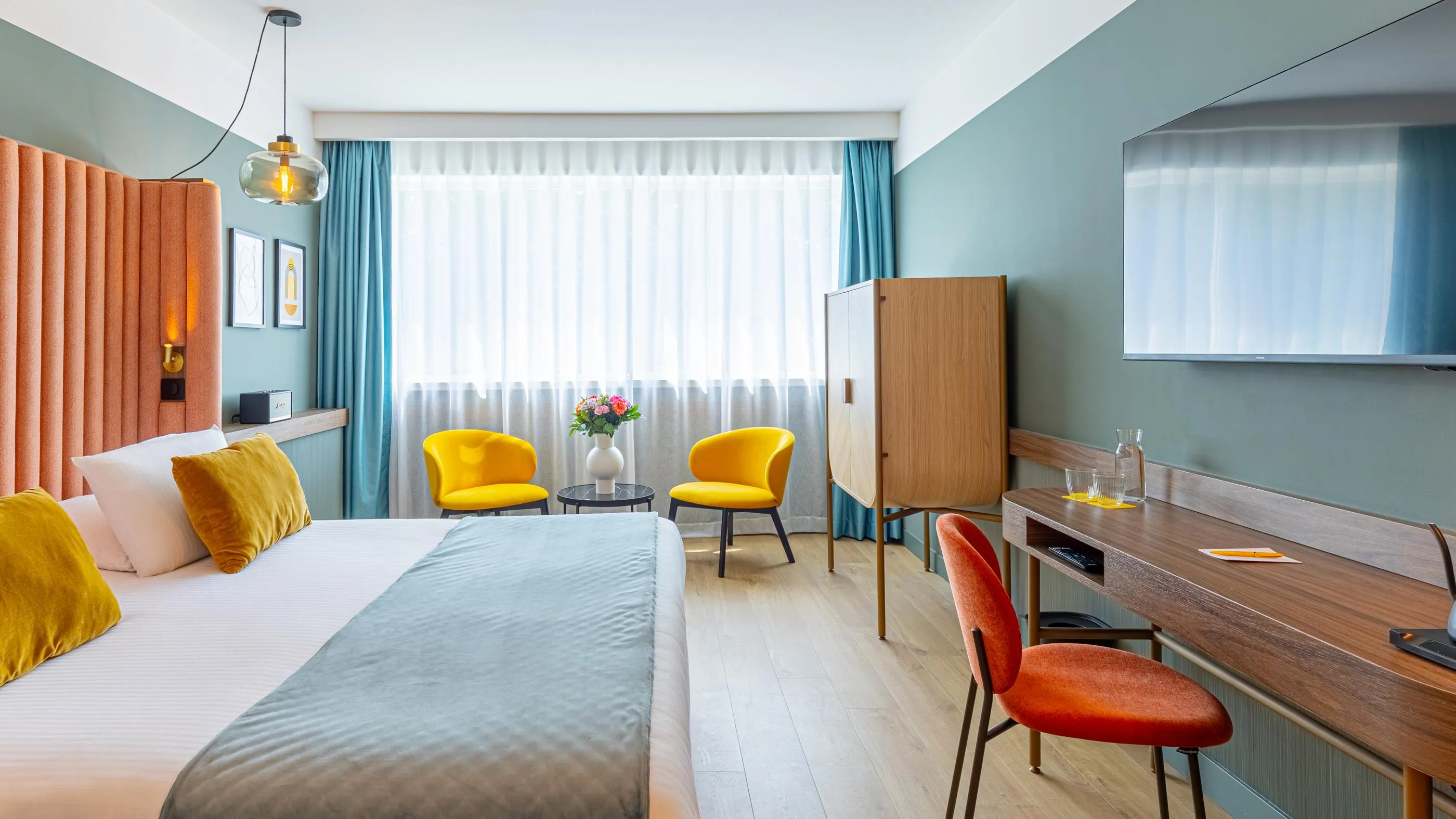 Zimmer mit Bett, zwei gelben Stühlen, Schreibtisch, Fernseher und großen Fenstern mit Vorhängen