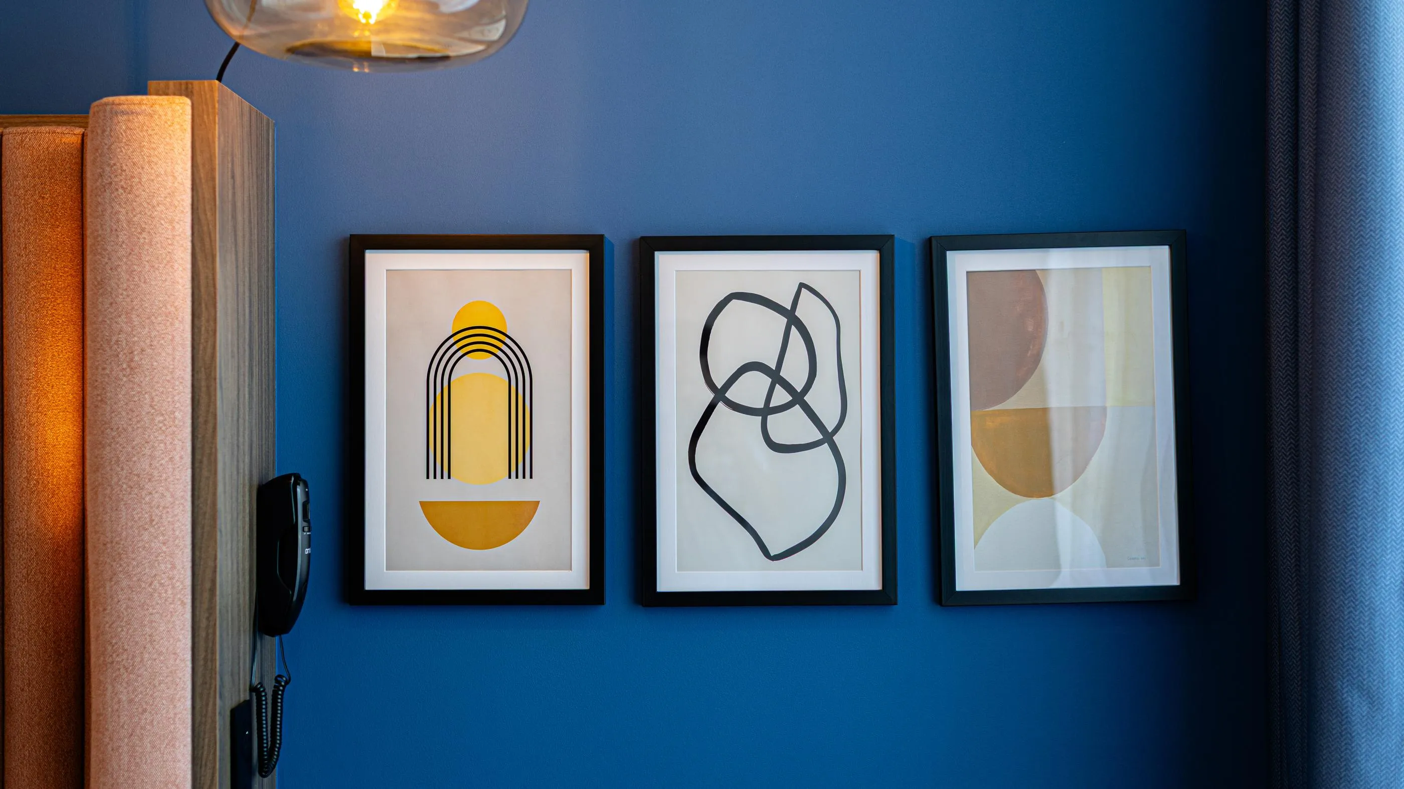 Trois cadres contenant des œuvres d'art abstraites sont accrochés sur un mur bleu près d'une lampe et d'un téléphone