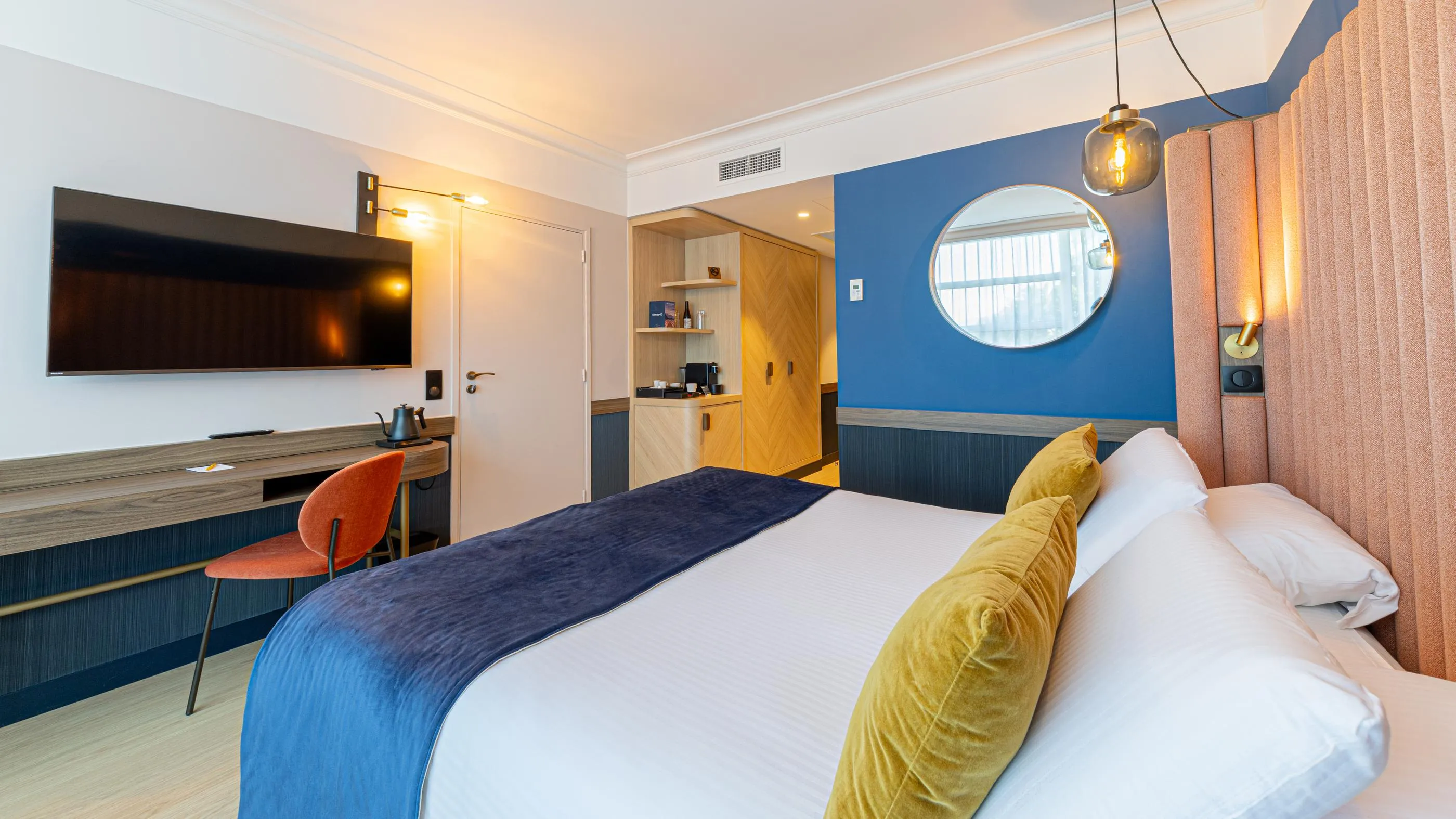 Chambre d'hôtel avec un lit double, une télévision, un bureau et une kitchenette visible à travers une porte ouverte