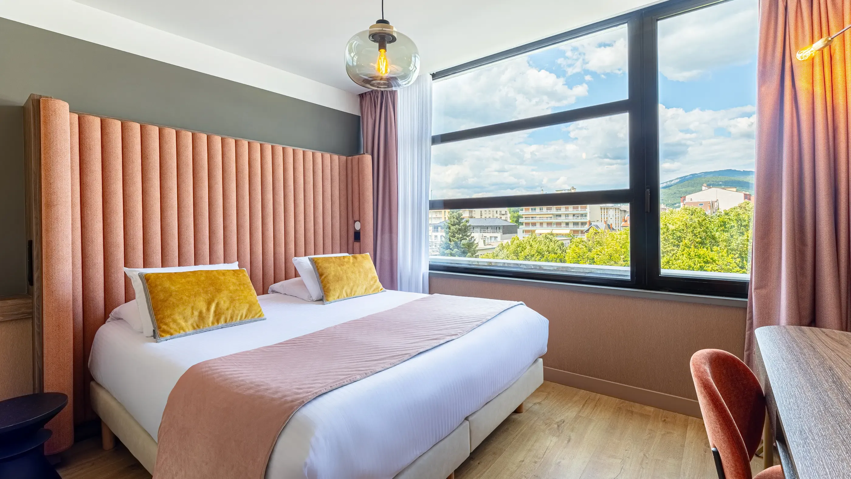 Zimmer mit einem Doppelbett mit weißen Laken und einer rosa Decke, zwei gelben Kissen, einem orangen Kopfteil und Blick auf Gebäude und Bäume durch ein großes Fenster.