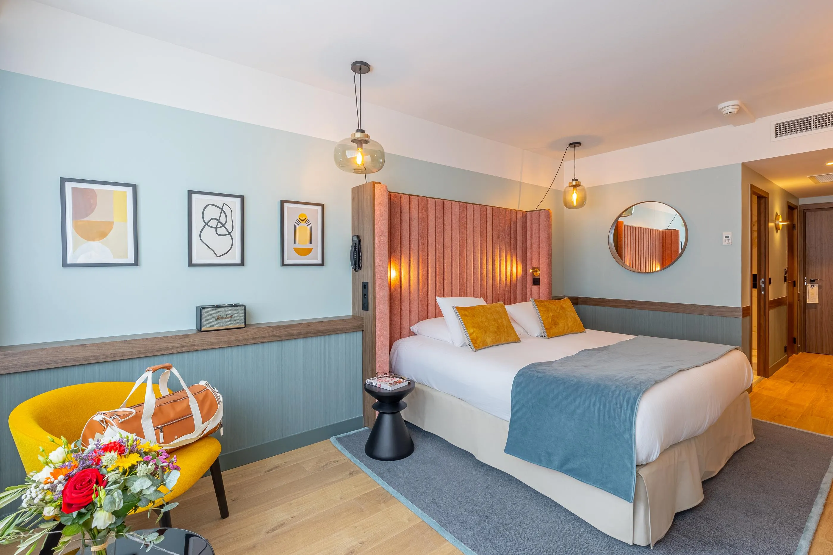 Zimmer mit einem Doppelbett, einem Stoffkopfteil, gelben Kissen, einer blauen Decke, einem schwarzen Nachttisch, einem gelben Sessel und Blumen auf dem Tisch.