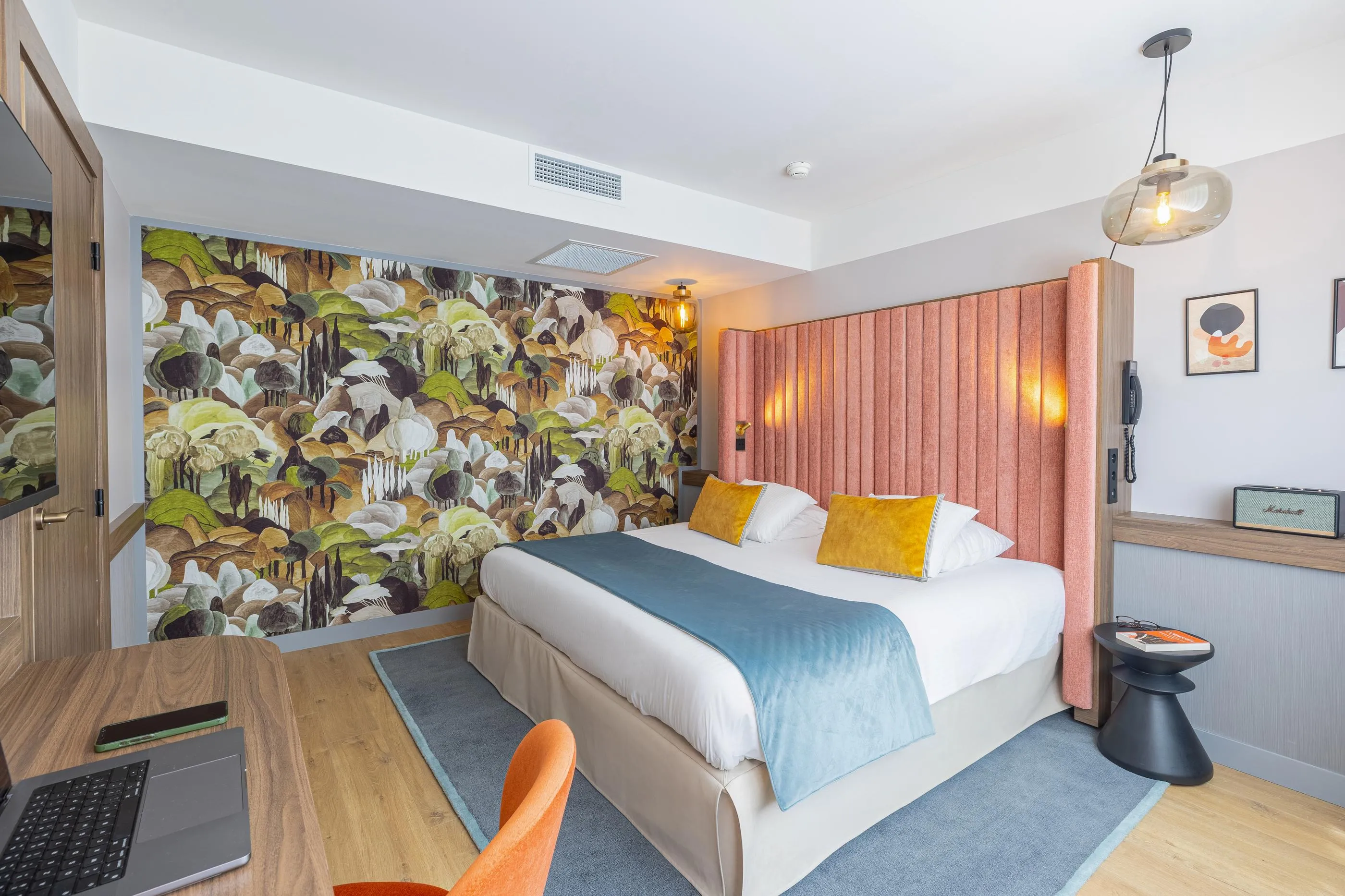 Chambre avec un lit, une table de travail, un fauteuil et un mur décoré de grandes feuilles et fleurs colorées