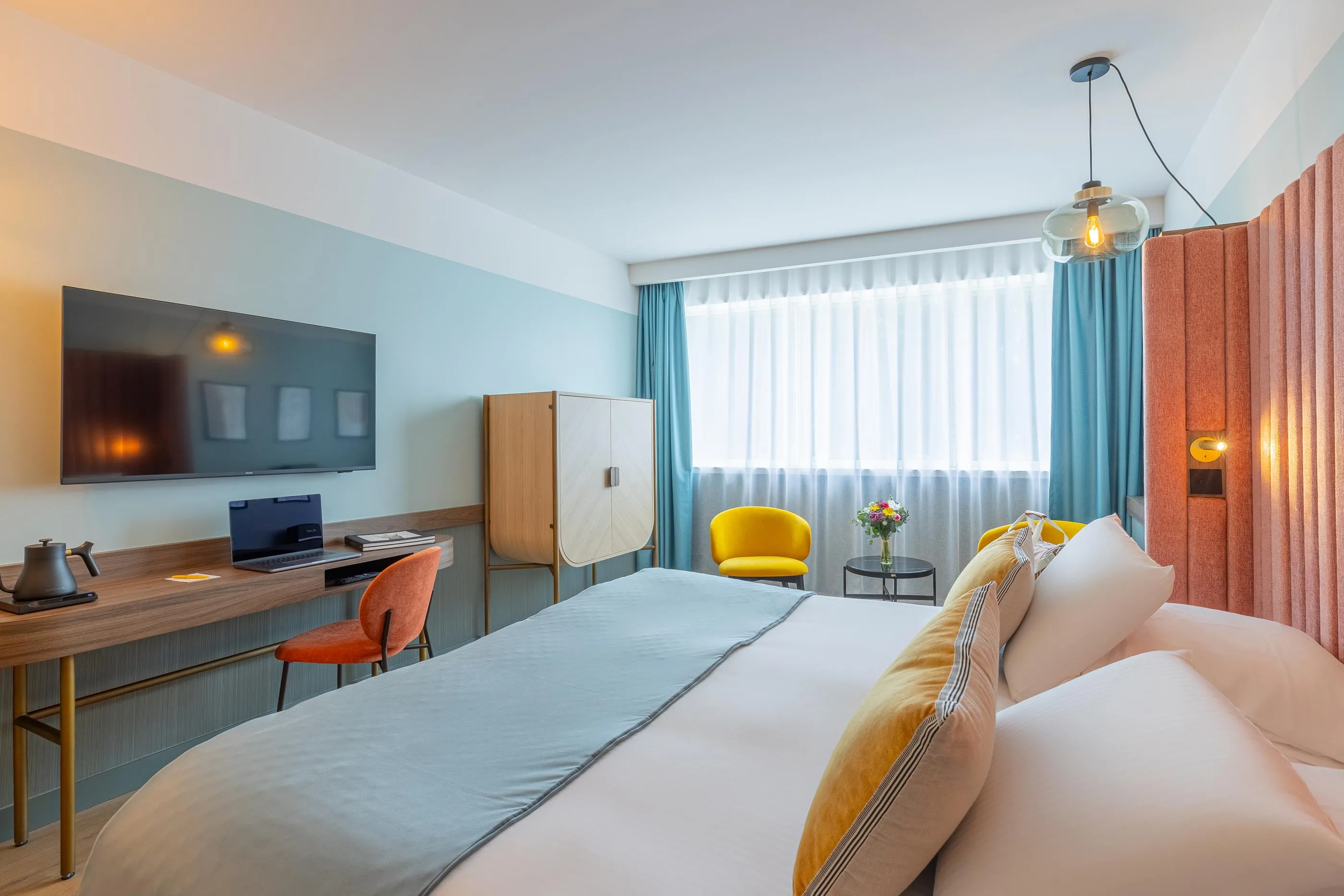Hotel room with a double bed, a television, a desk, a yellow chair, and a window with curtains.