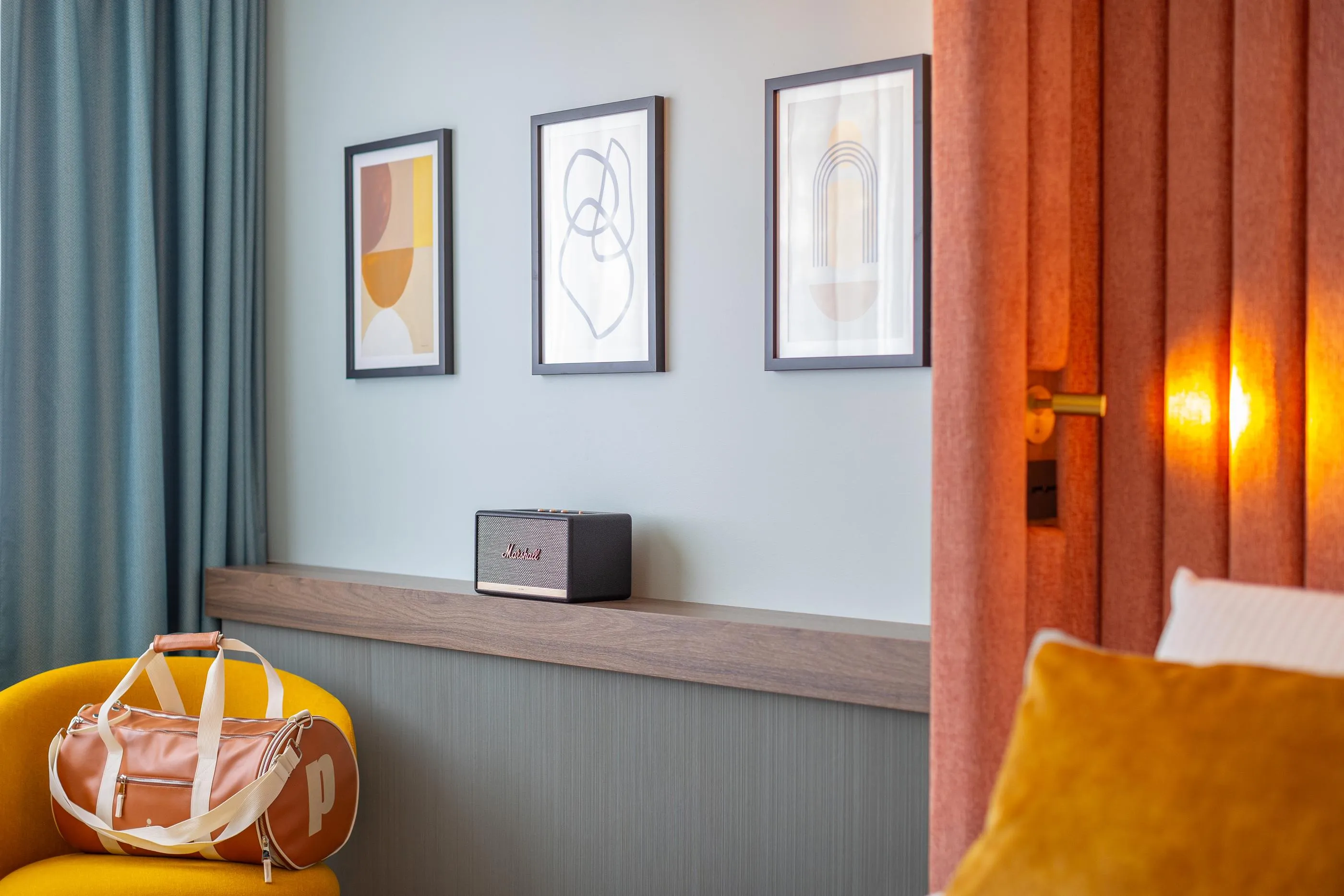 Ein Salon mit gerahmten Kunstwerken an der Wand, einem Lautsprecher auf einem Regal, einer Ledertasche auf einem gelben Sessel und einer halb geöffneten Tür mit Licht, das durch die Vorhänge fällt.