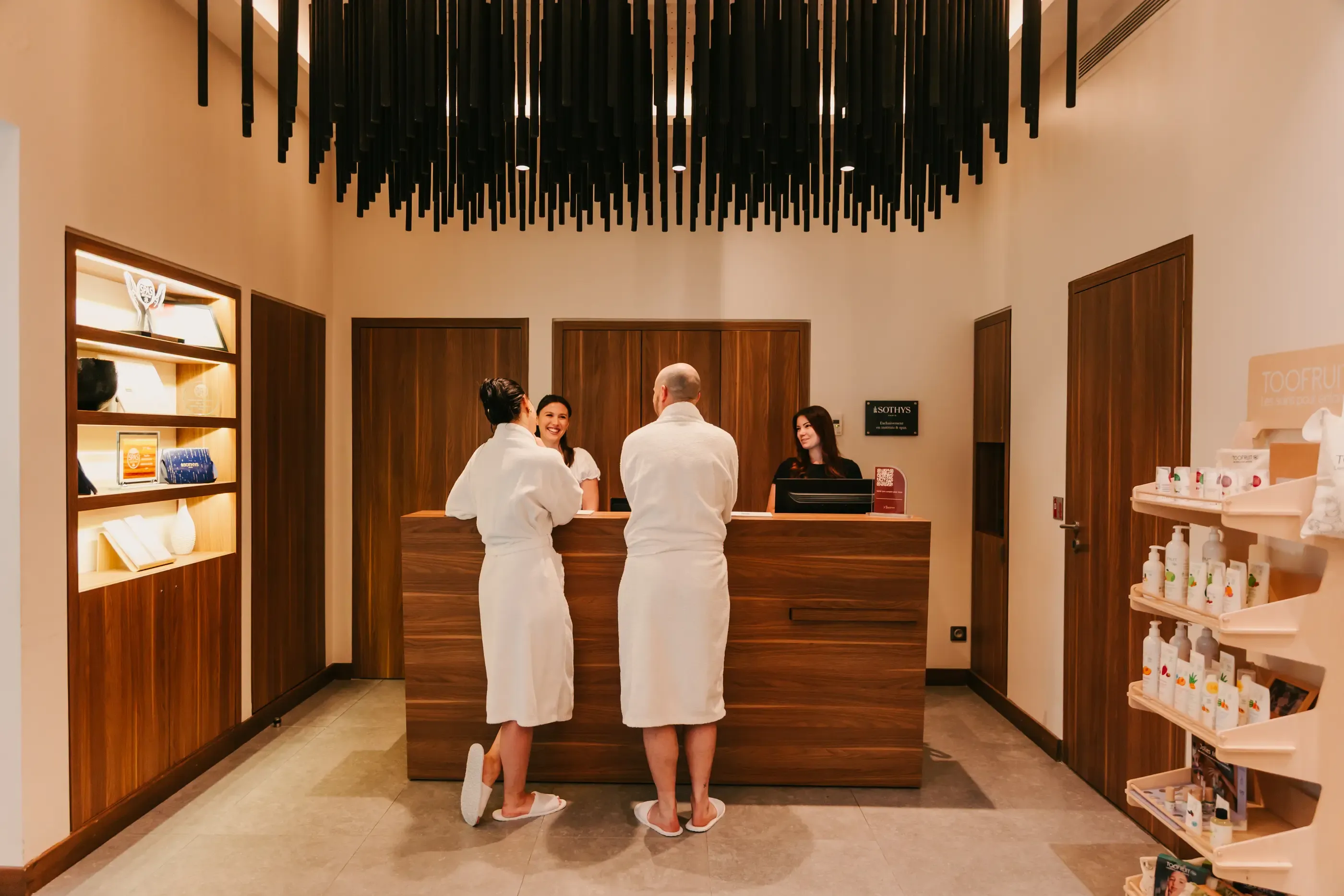 Deux personnes en robe de bain blanche discutent avec une réceptionniste derrière un comptoir en bois dans un spa, avec des produits de soins sur une étagère à droite