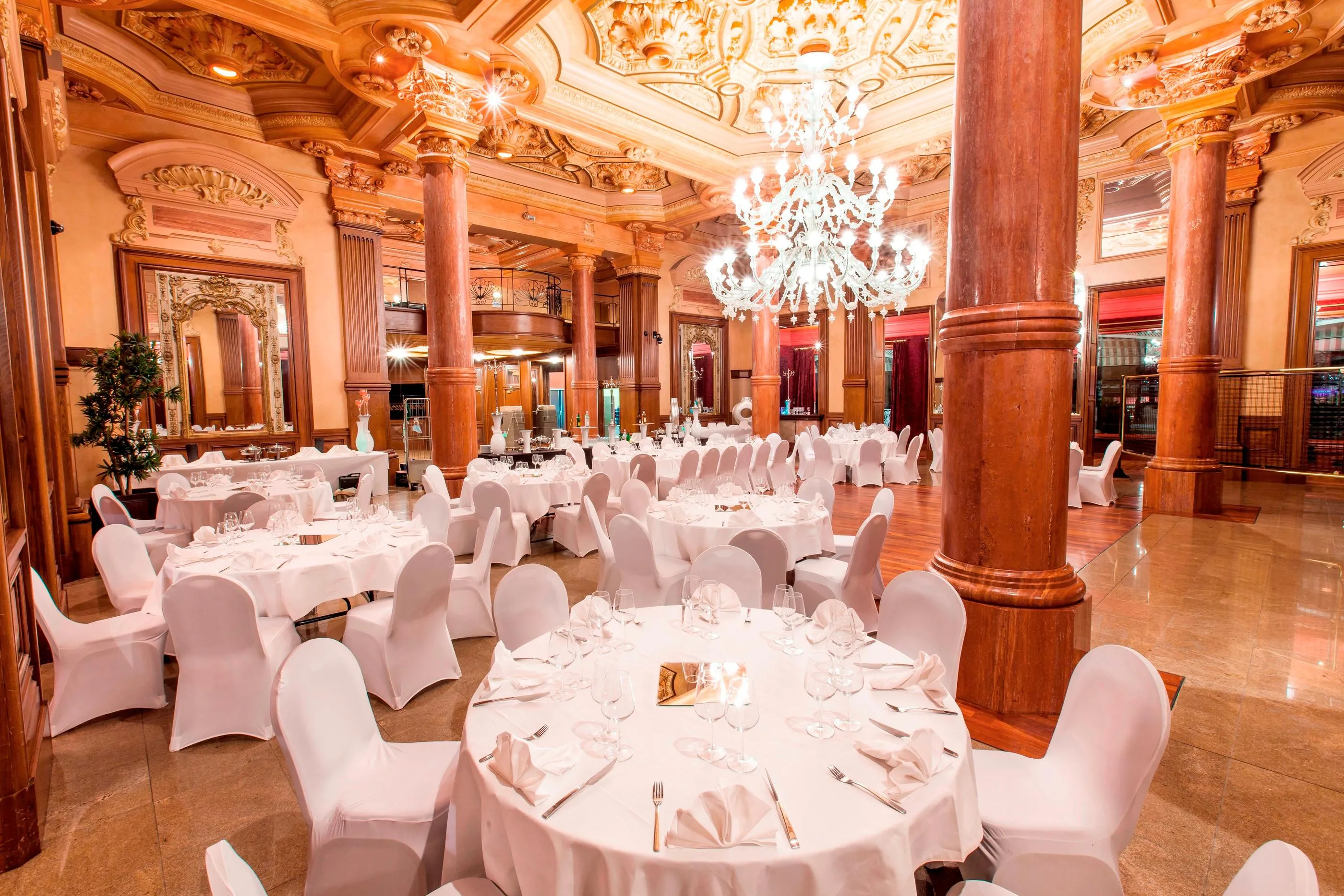 Salle de réception luxueuse avec tables rondes couvertes de nappes blanches, chaises assorties et lustres suspendus au plafond orné
