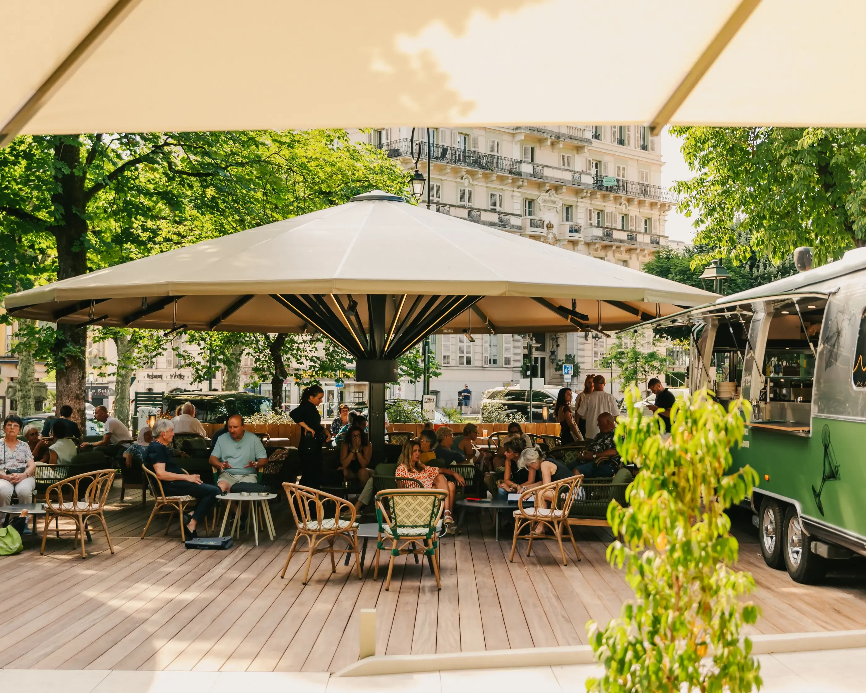 Une terrasse de café en plein air avec des clients assis sous des parasols, entourée d'arbres et de bâtiments anciens