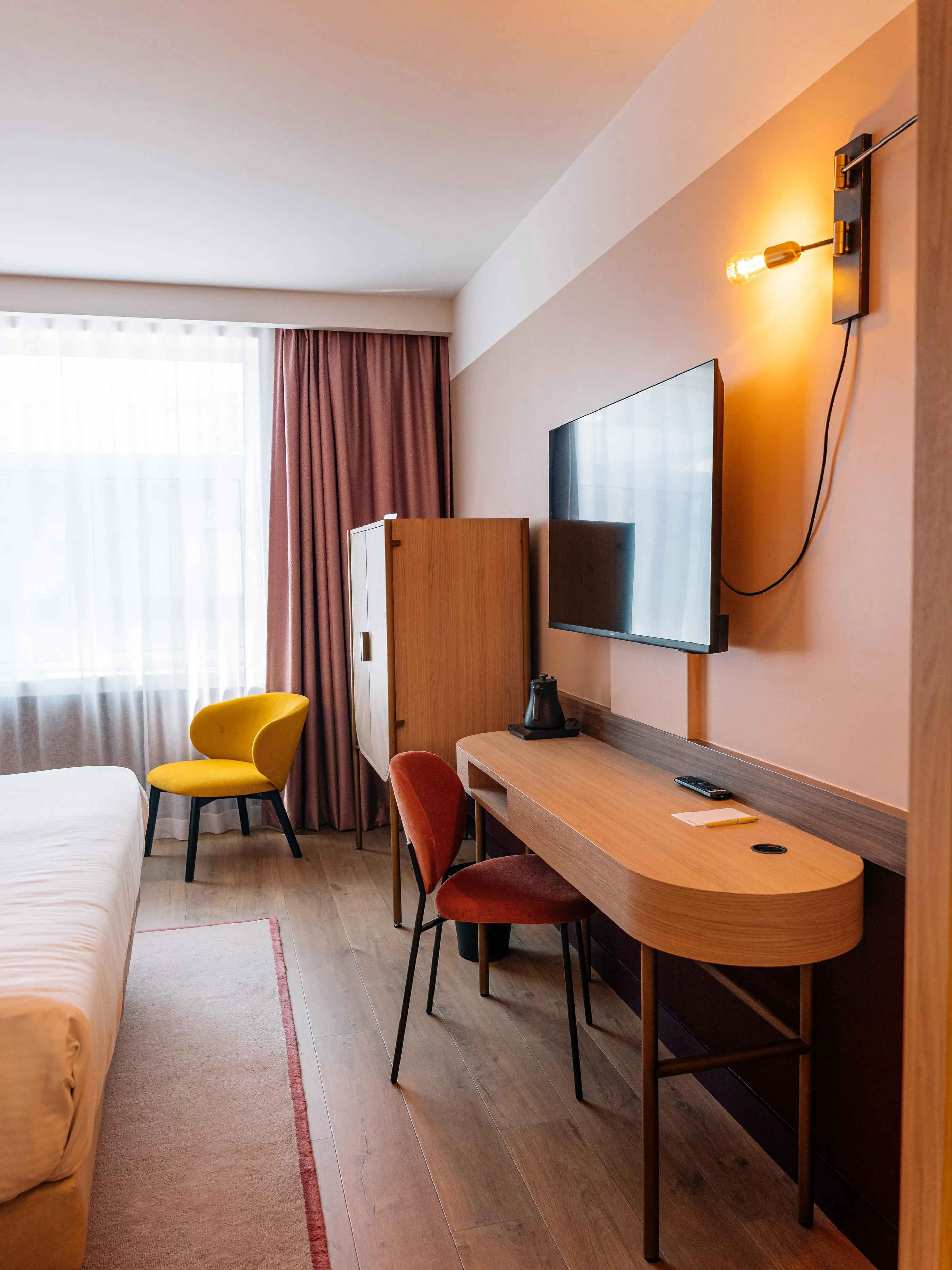 Zimmer mit einem Bett, einem Schreibtisch, einem Fernseher, einem gelben Sessel und einem Fenster mit Vorhängen