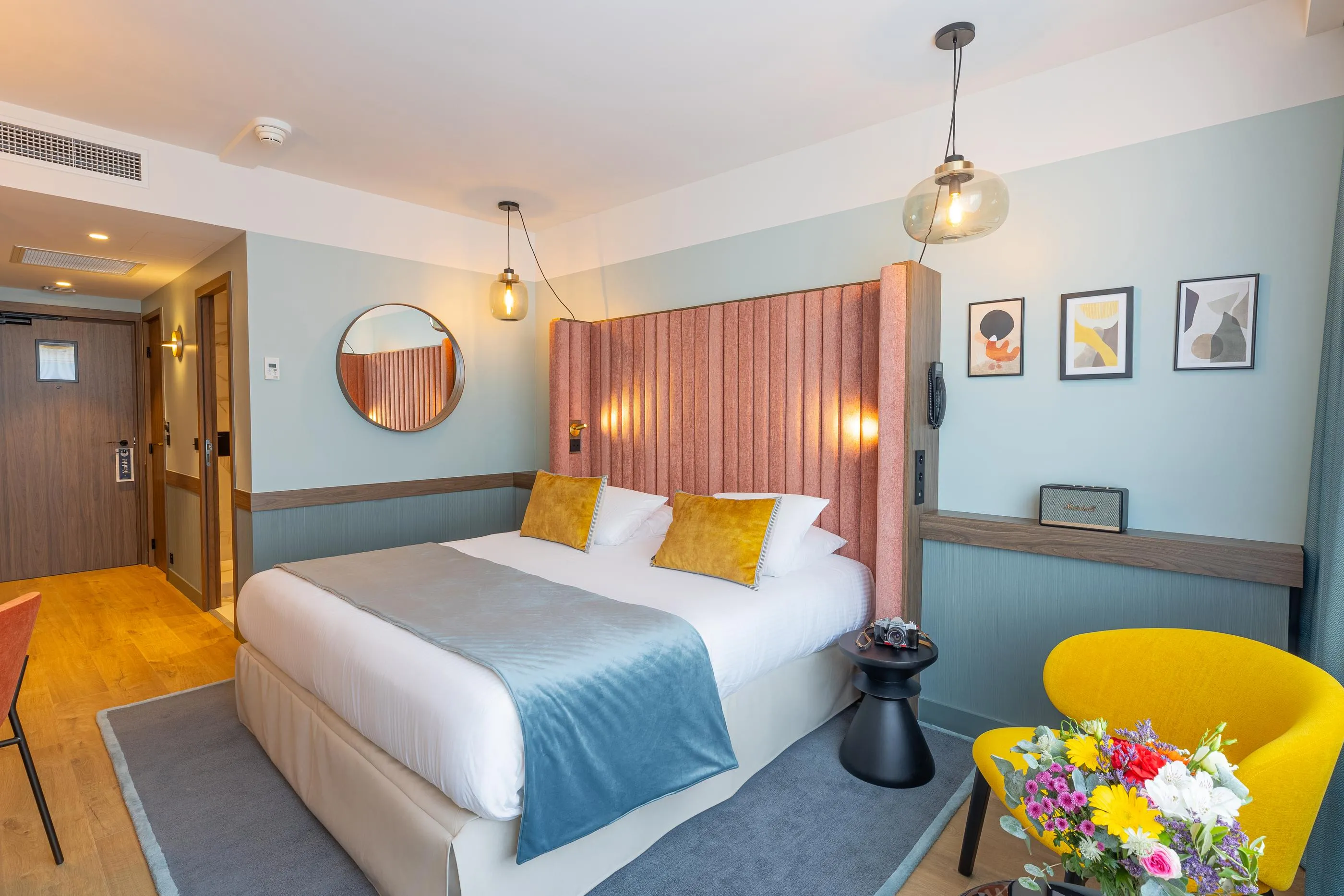 Room with a double bed, a fabric headboard, yellow cushions, a bedside table, a yellow armchair, and flowers on the table.
