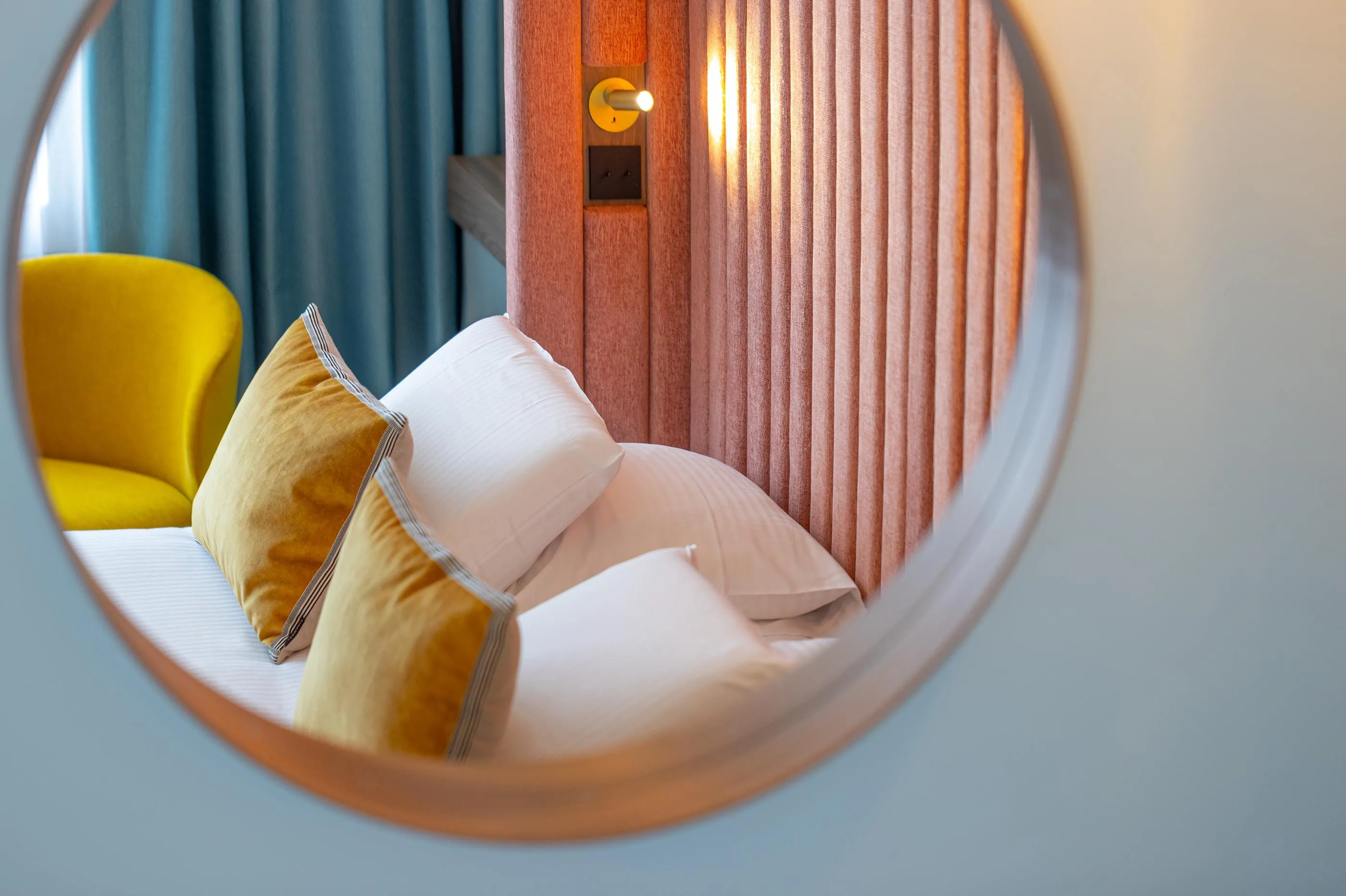 Reflet dans un miroir rond d'un lit avec des coussins beiges et blancs et d'une chaise jaune près de rideaux gris et d'une porte en bois