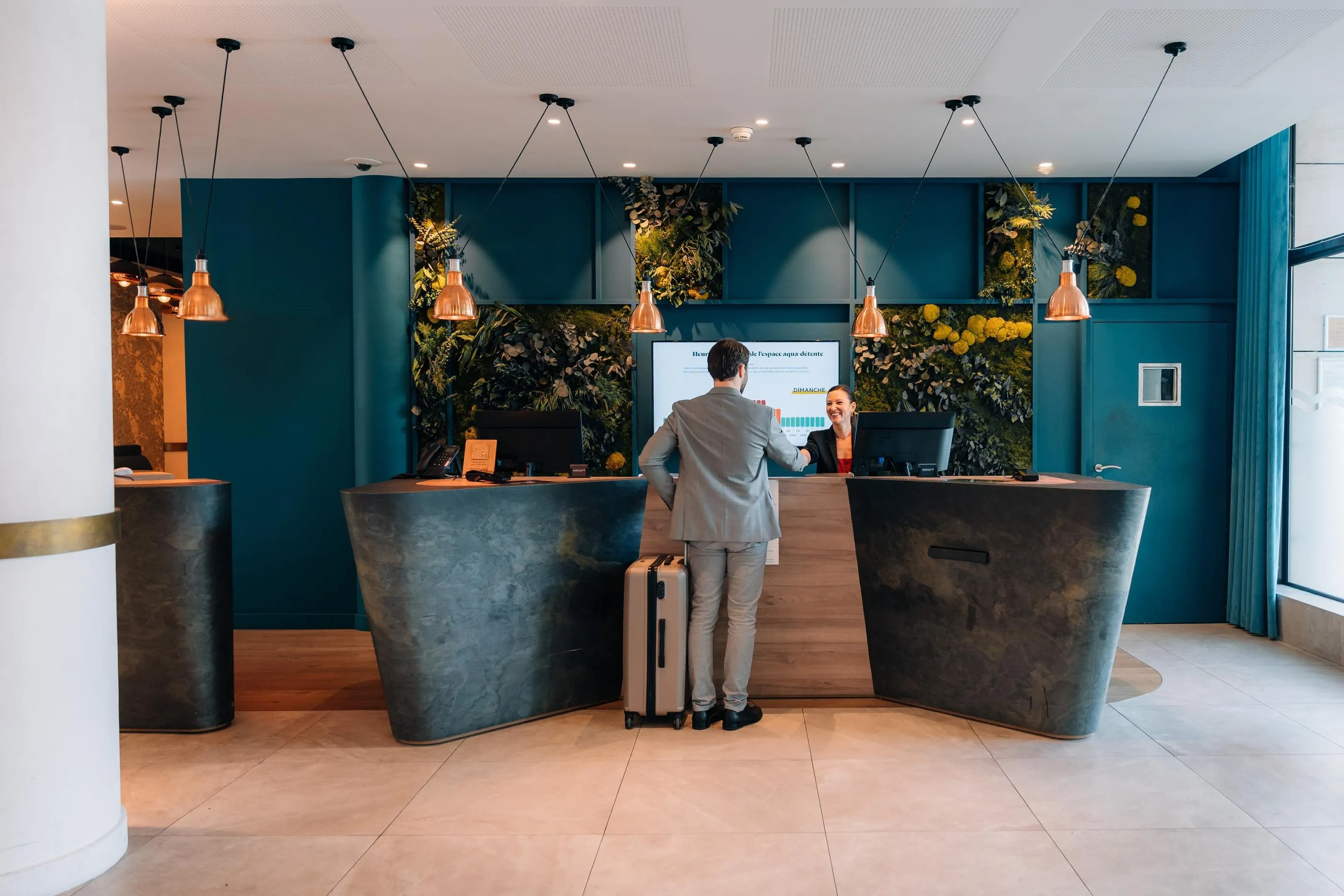 Un homme en costume serre la main d'une réceptionniste derrière un comptoir dans un hall d'hôtel moderne avec des plantes murales