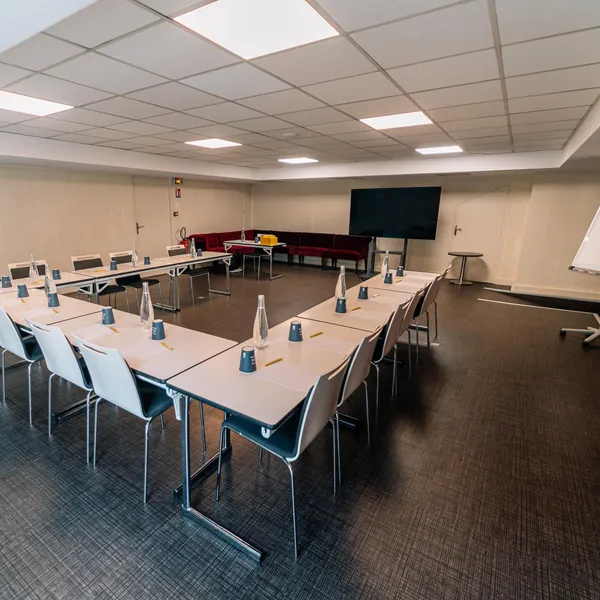 Salle de réunion avec tables disposées en U, chaises, écran, tableau blanc et rangée de sièges rouges