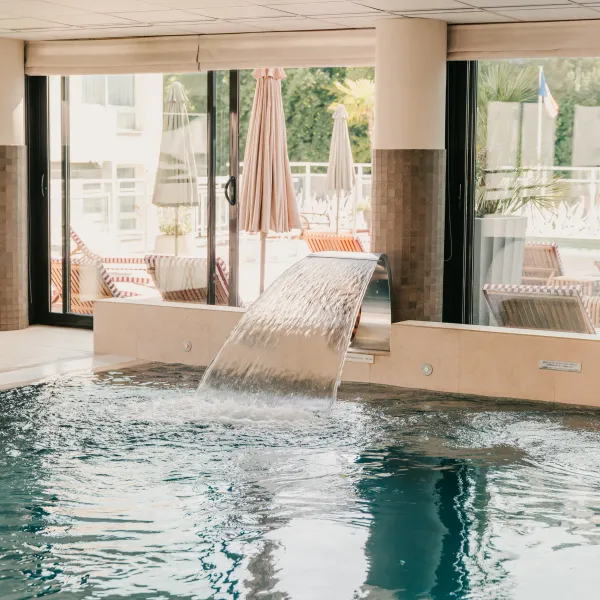 Innenpool mit kleinem Becken und Wasserfall, umgeben von großen Glasfronten, die auf eine Terrasse mit Liegestühlen führen