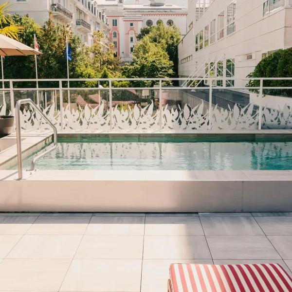Rechteckiges Schwimmbecken mit Treppen und Handlauf, umgeben von hellem Fliesenboden, Liegestühlen und Topfpflanzen, mit einem Gebäude im Hintergrund