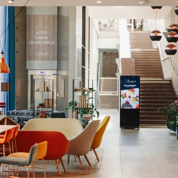 Moderner Eingangsbereich mit Treppe, farbigen Sitzgelegenheiten, Pflanzen und Anschlagtafeln, die den Zugang zum Casino und die Frühstückszeiten anzeigen