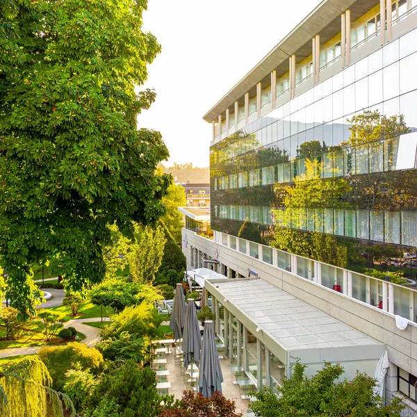 Un bâtiment moderne avec de grandes fenêtres reflétant des arbres verts, adjacent à une terrasse extérieure avec des tables et des chaises