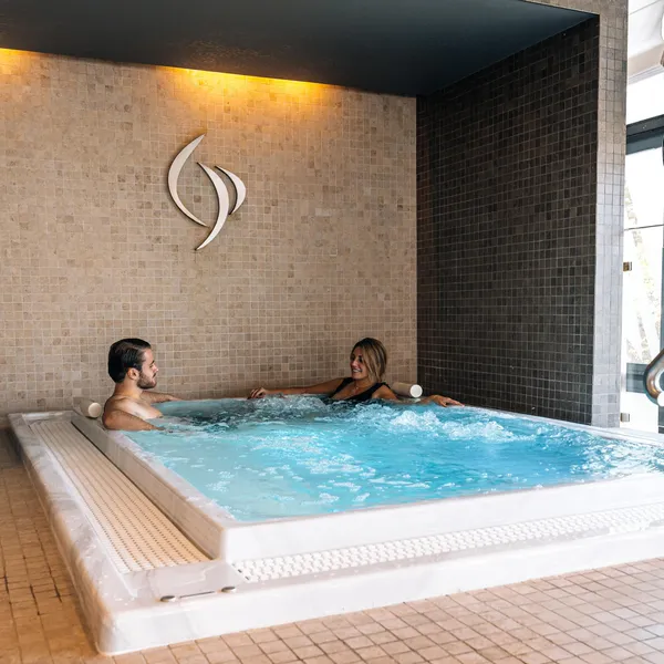 Deux personnes sont assises dans un jacuzzi, entourées de carrelage, avec une grande fenêtre à proximité