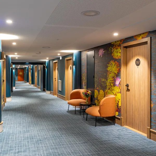 Couloir d'hôtel avec portes en bois, murs décorés de motifs colorés et sièges orange près d'une table basse