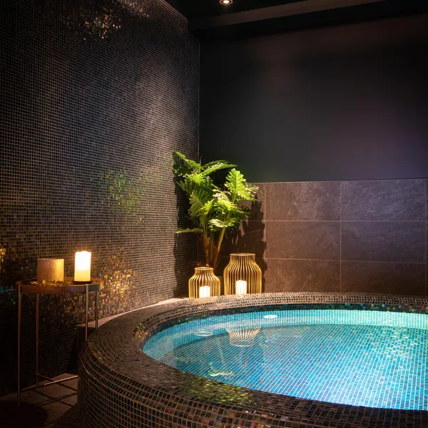 Innenjacuzzi mit blauer Beleuchtung, umgeben von Mosaiken und Pflanzen, mit Kerzen und einer Lampe in der Nähe