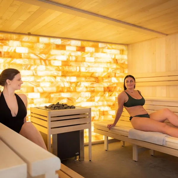 Deux femmes assises dans un sauna en bois éclairé par une paroi lumineuse, l'une en maillot noir et l'autre en tenue verte