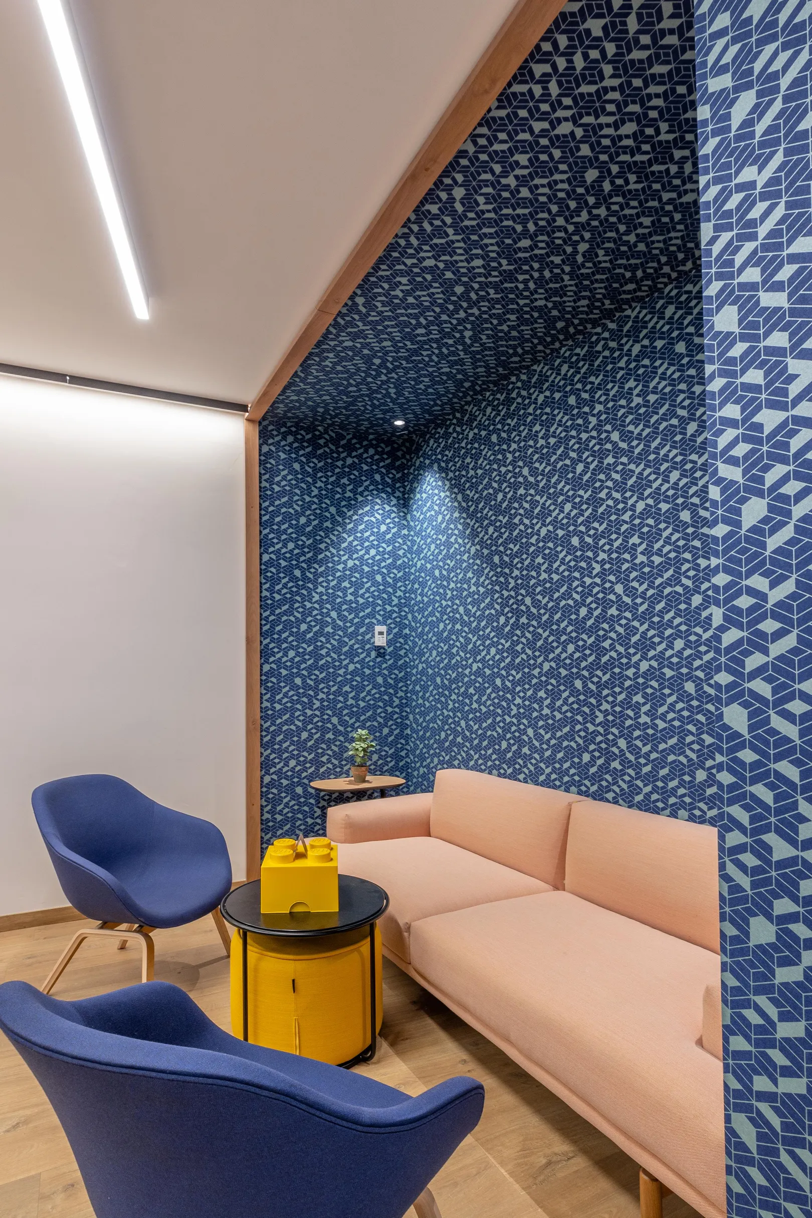 Ein Wohnzimmer mit einem beigen Sofa, zwei blauen Sesseln und einem gelben Couchtisch, vor einer mit blauen und grauen geometrischen Mustern verzierten Wand.