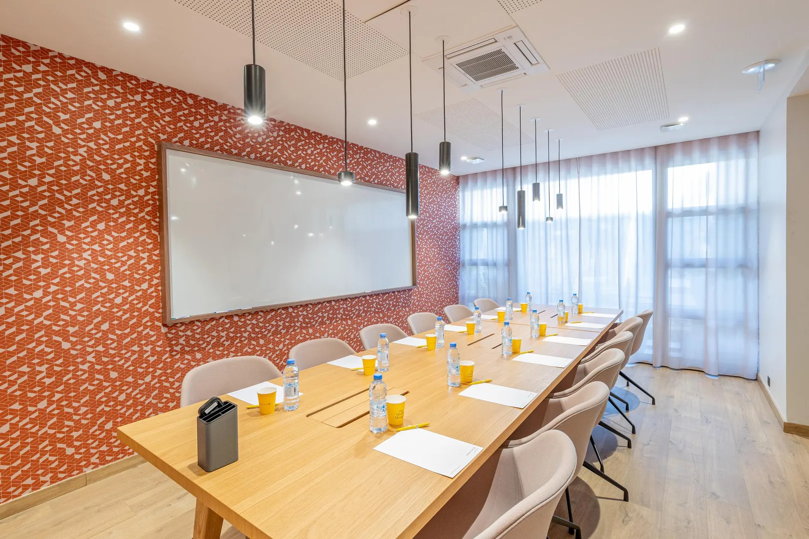 Besprechungsraum mit einem Holztisch, Stühlen, Wasserflaschen, Bechern und einer Whiteboard-Wand an einer mit roten geometrischen Mustern verzierten Wand