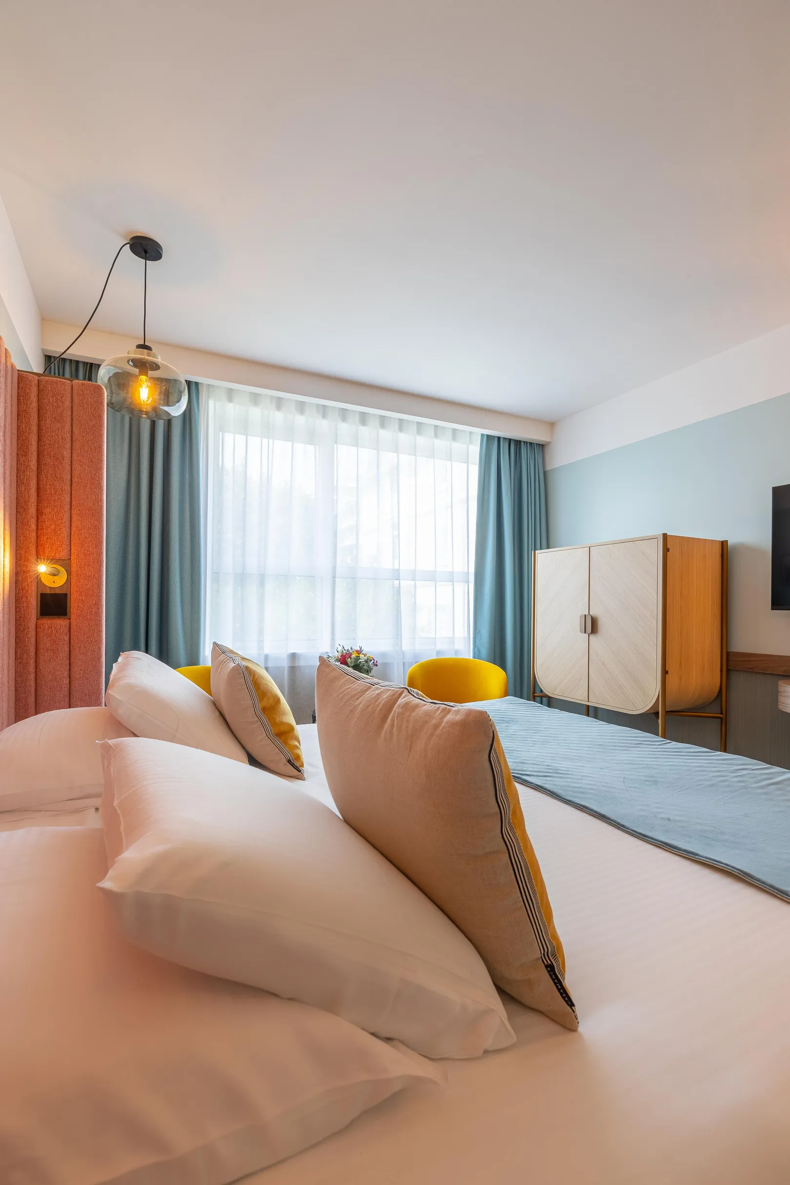 Zimmer mit einem Bett mit weißen Decken und bunten Kissen, einem Fenster mit Vorhängen, einer Wandlampe und einem Holzmöbel