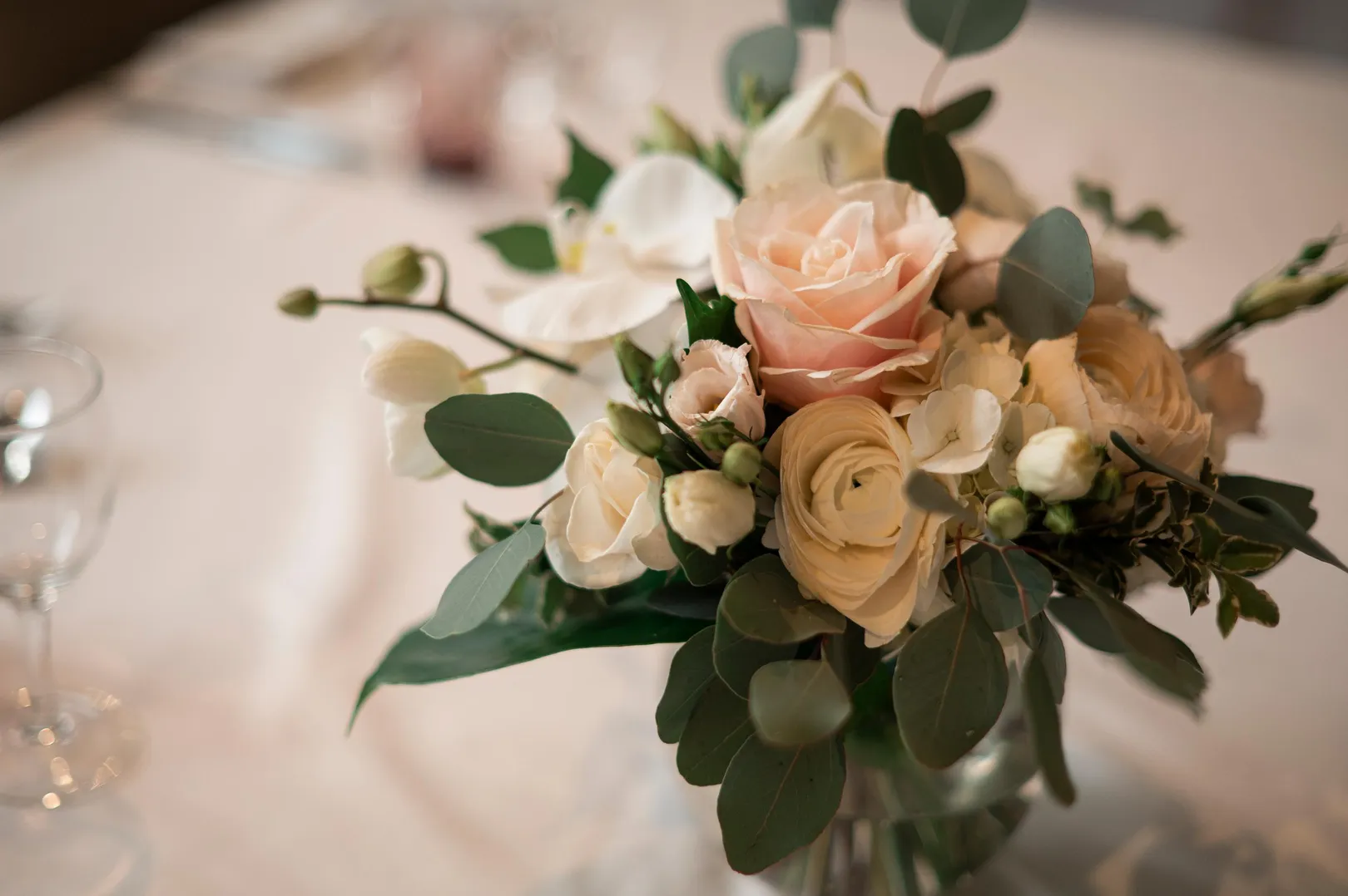 Strauß aus weißen und rosa Rosen mit grünem Blattwerk auf einem weißen Tisch mit einem Weinglas im Hintergrund