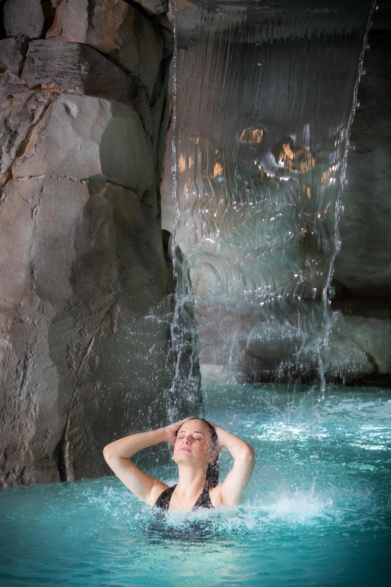 Eine Person ist in einem Pool unter einem Wasserfall eingetaucht, die Arme auf dem Beckenrand abgelegt.