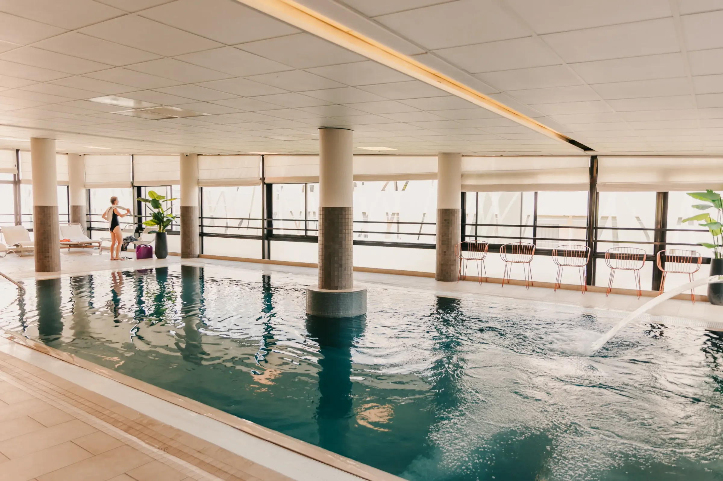Piscine intérieure avec une personne en maillot de bain près de chaises longues et de tables, entourée de grandes fenêtres et de colonnes