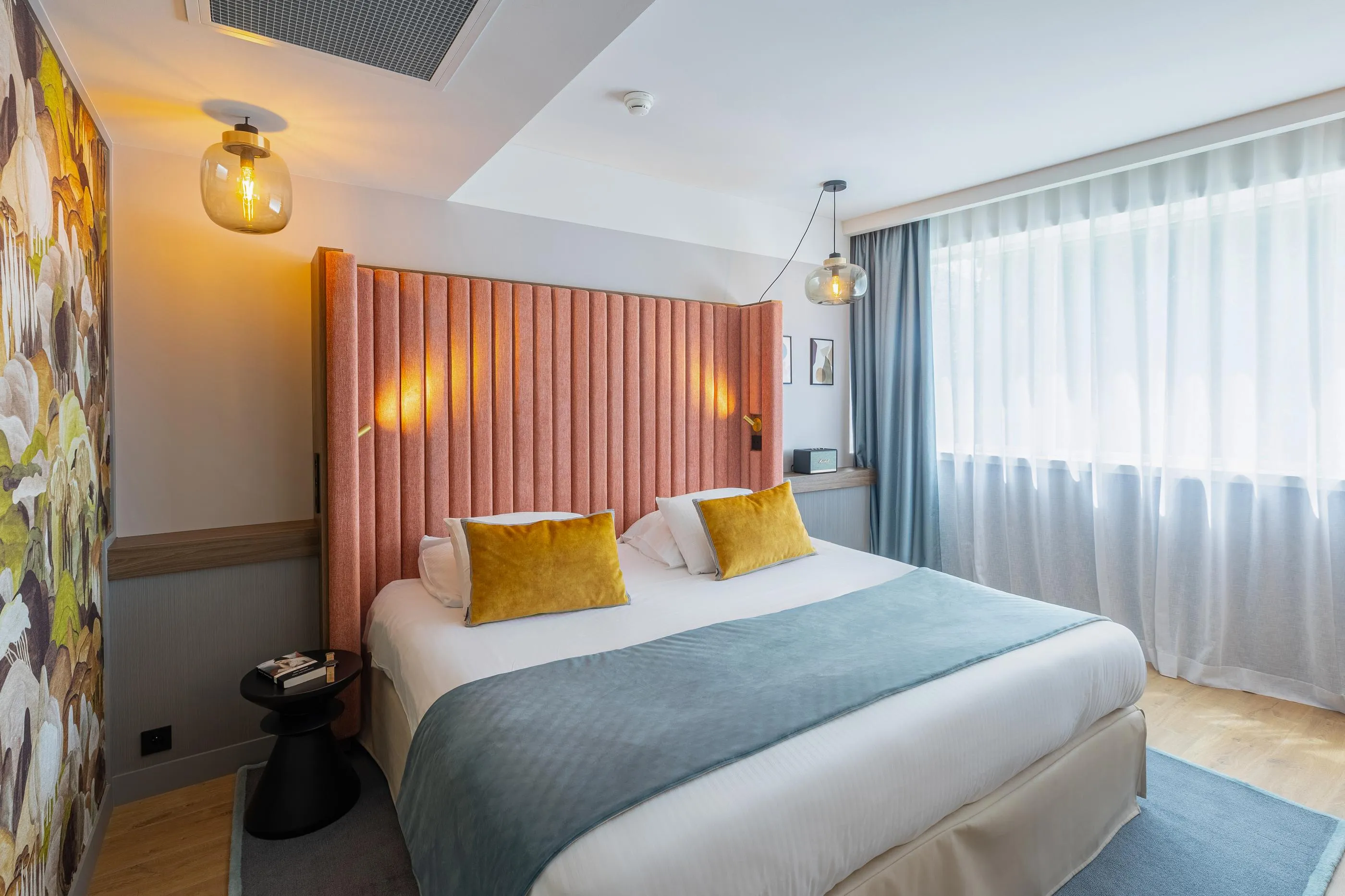 Room with a double bed covered by a blue blanket and yellow pillows, white and gray curtains, wall lamps, and floral wall decoration.