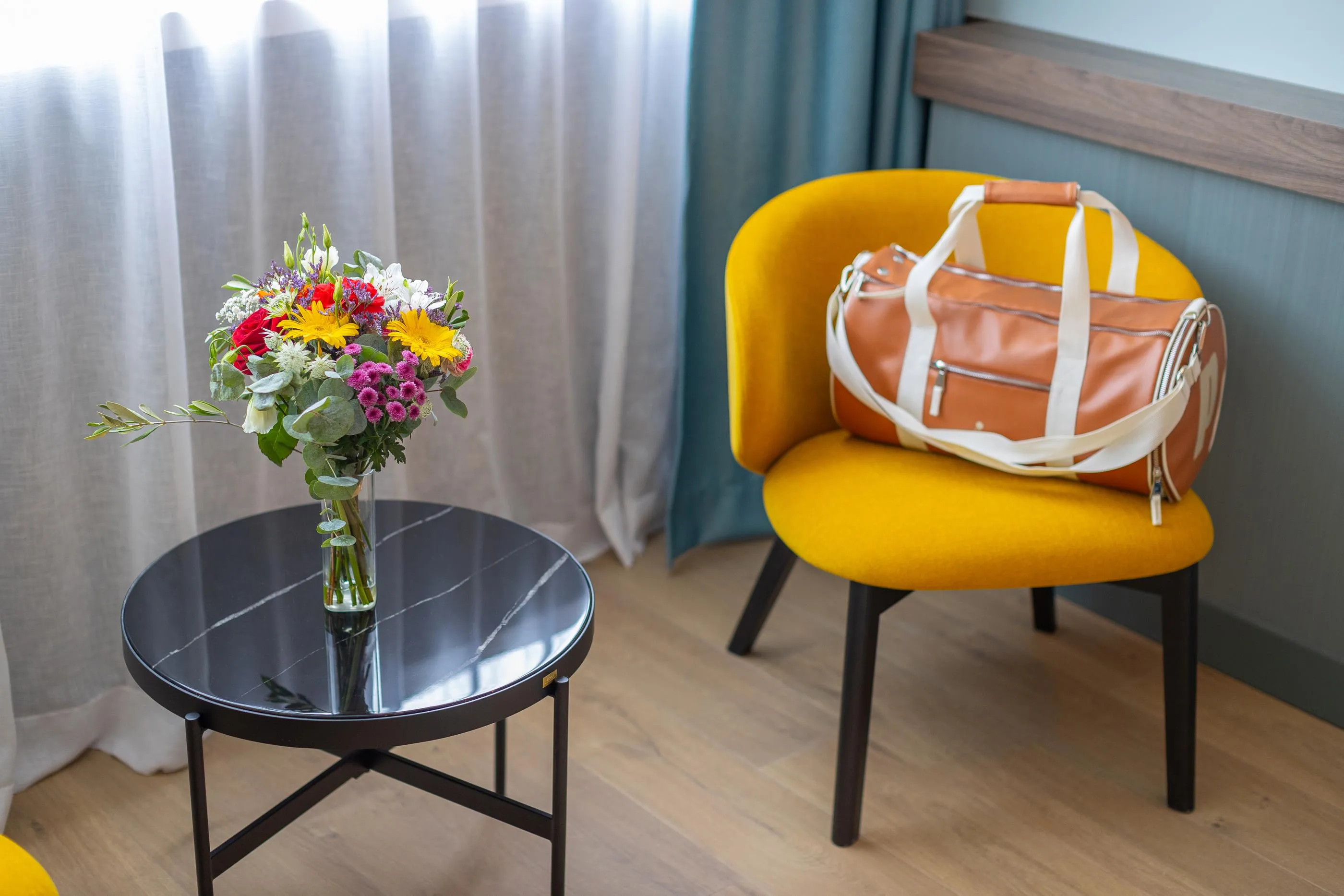 A black round table with a vase of colorful flowers next to a yellow chair with a leather bag on it.
