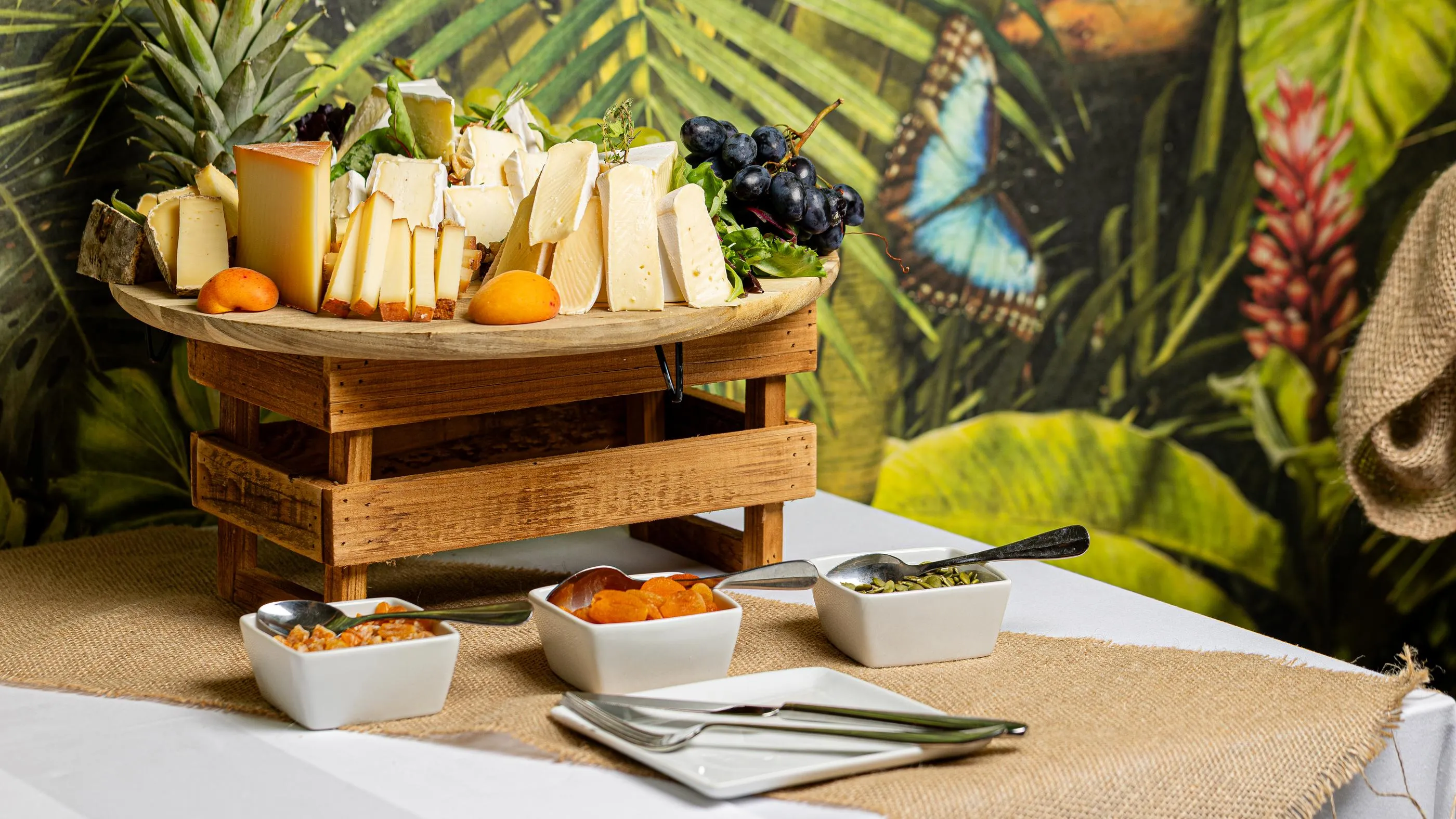 A cheese and fruit platter with three small bowls of accompaniments is placed on a table covered with a jute tablecloth in front of a tropical vegetation backdrop.