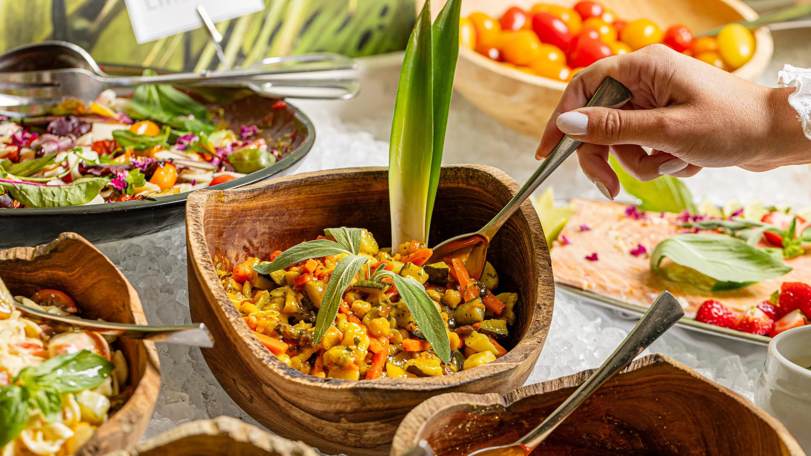 A person serves a vegetable dish in a wooden bowl on a buffet table with various other dishes.