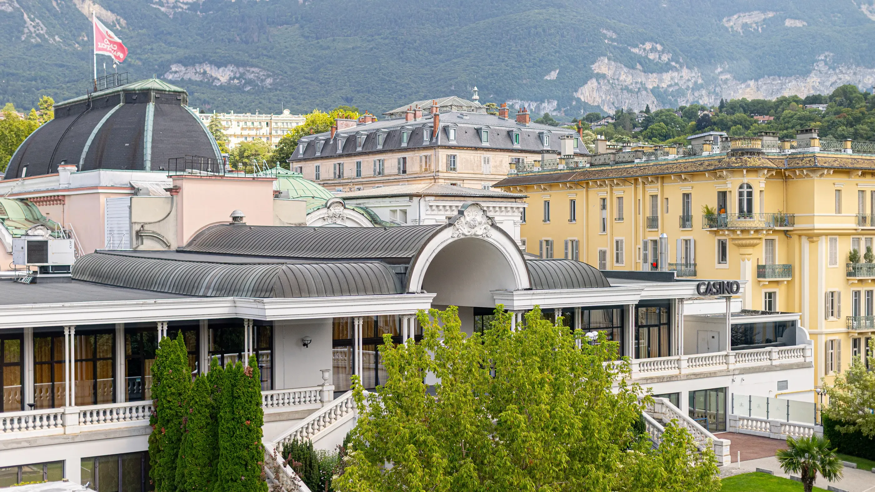 A casino with classic architecture and metal roofs, surrounded by historic buildings and mountains in the background.