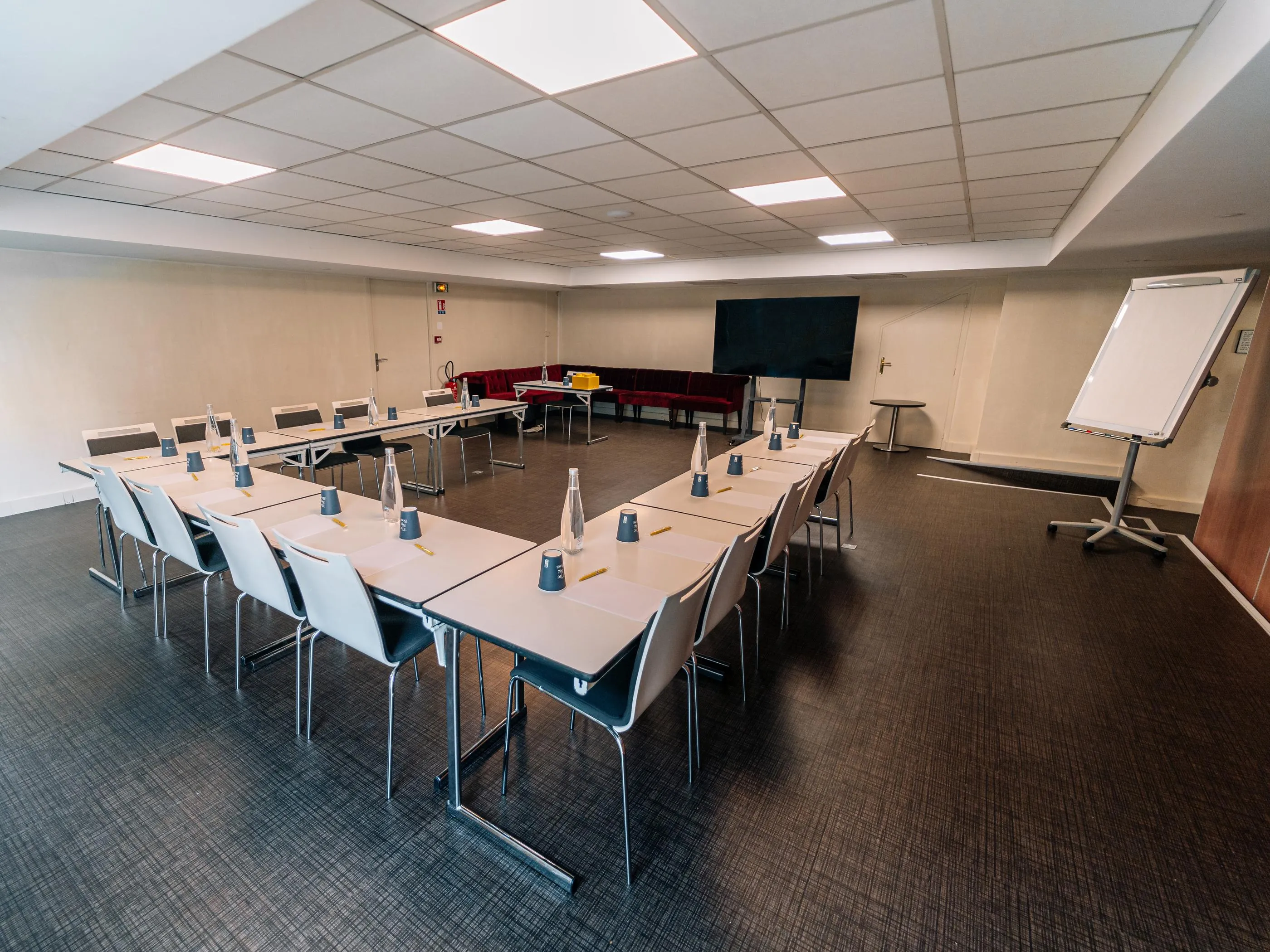 Besprechungsraum mit U-förmig angeordneten Tischen, Stühlen, Bildschirm, Whiteboard und einer Reihe roter Sitzplätze