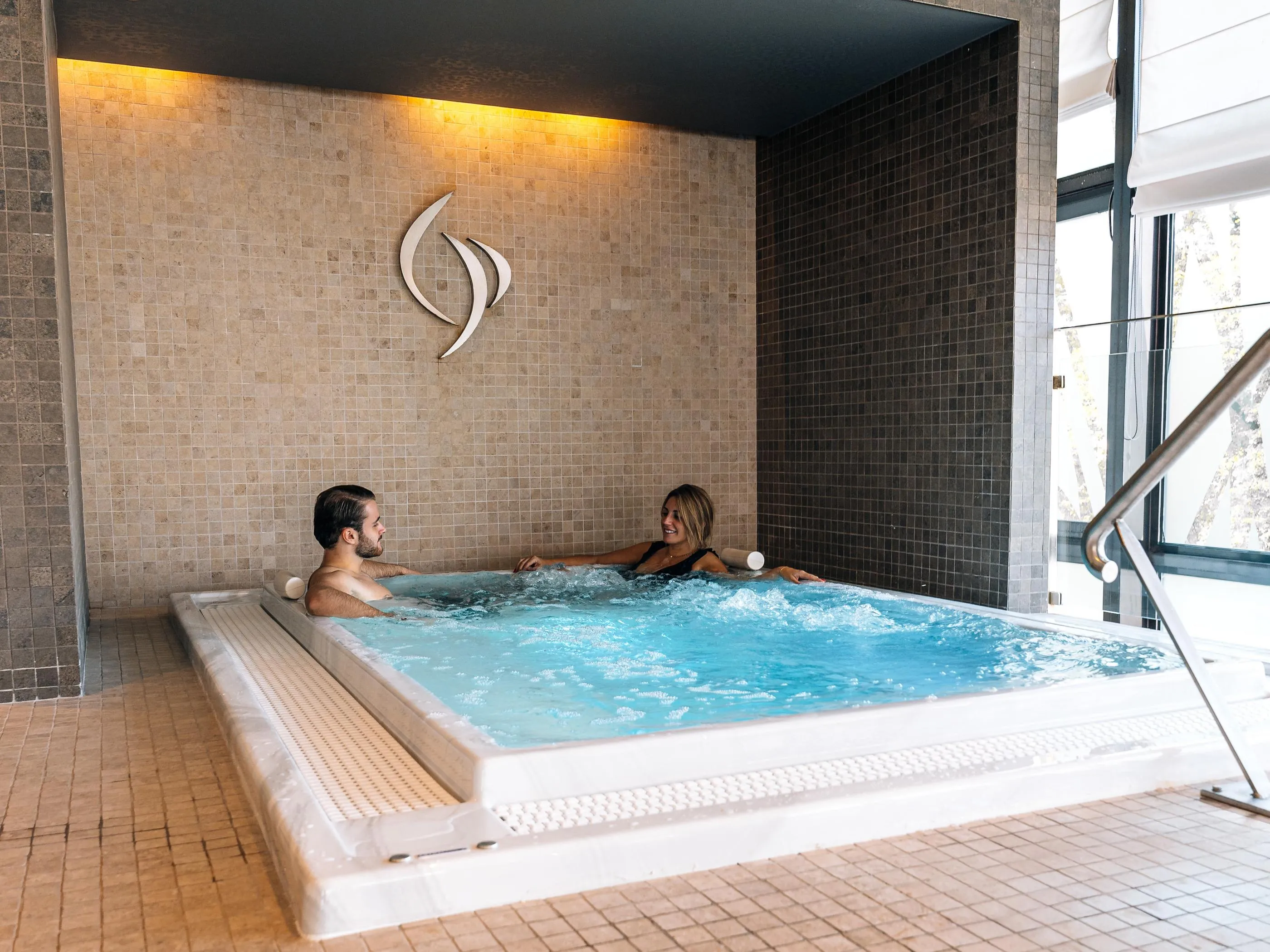 Two people are sitting in a jacuzzi, surrounded by tiles, with a large window nearby.