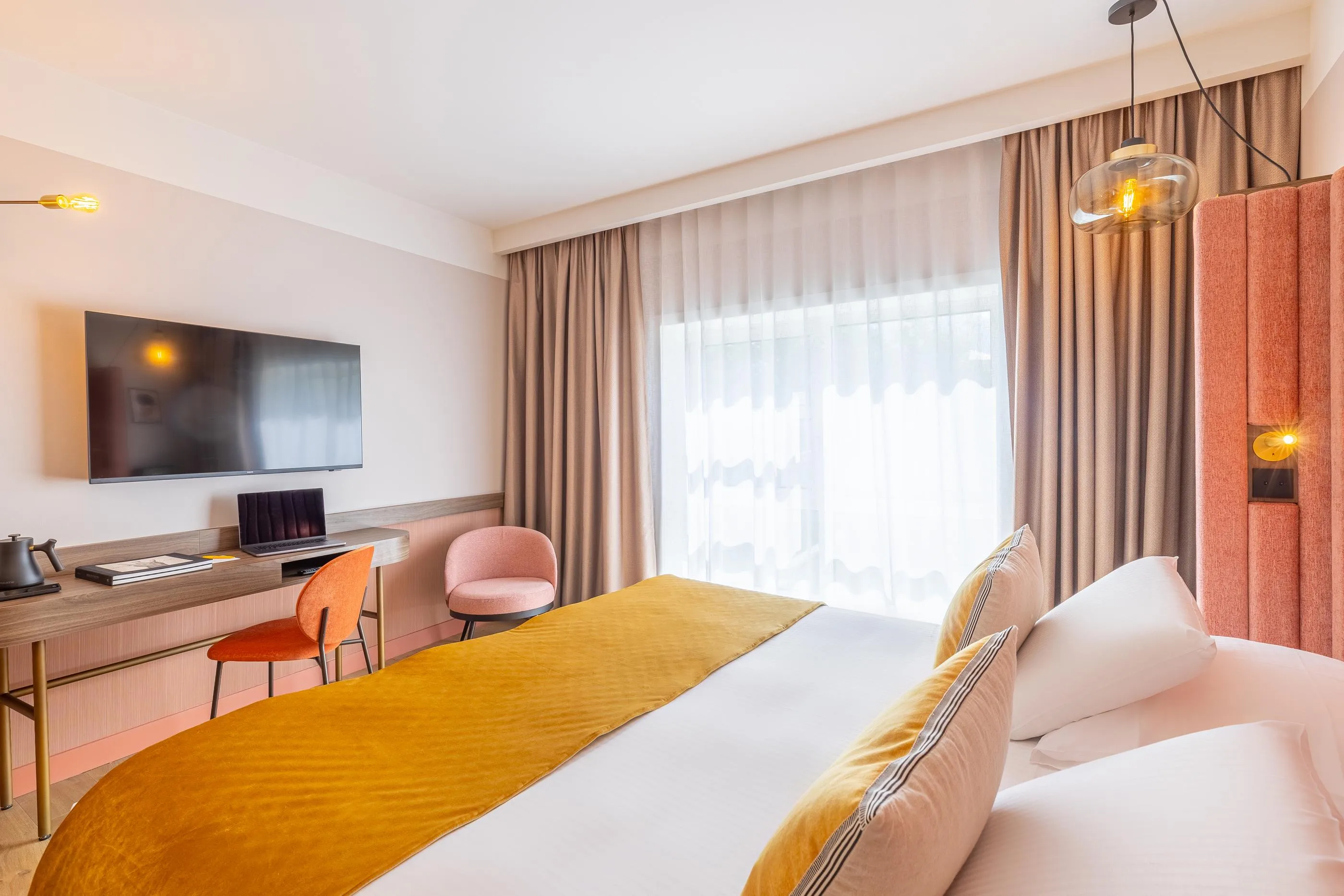 Hotelzimmer mit einem Doppelbett, einem Schreibtisch, einem Wandfernseher und einem großen Fenster mit Vorhängen