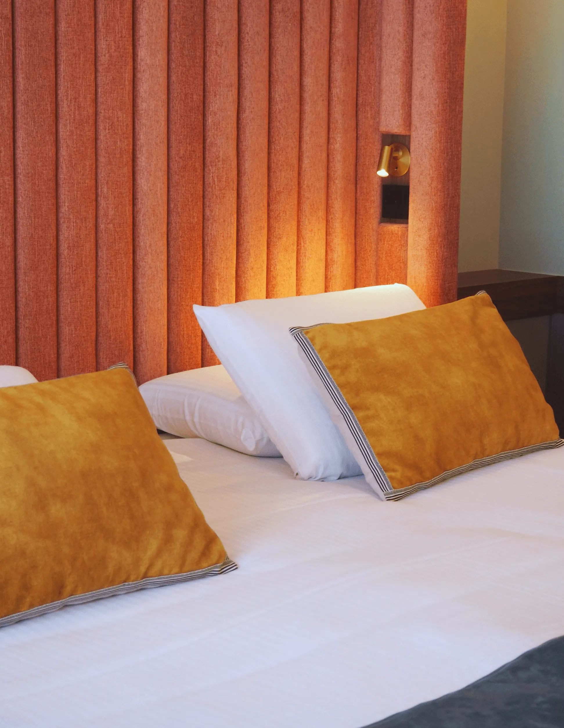 Bed with white sheets and orange and white pillows, a wall with orange curtains and a lit bedside lamp