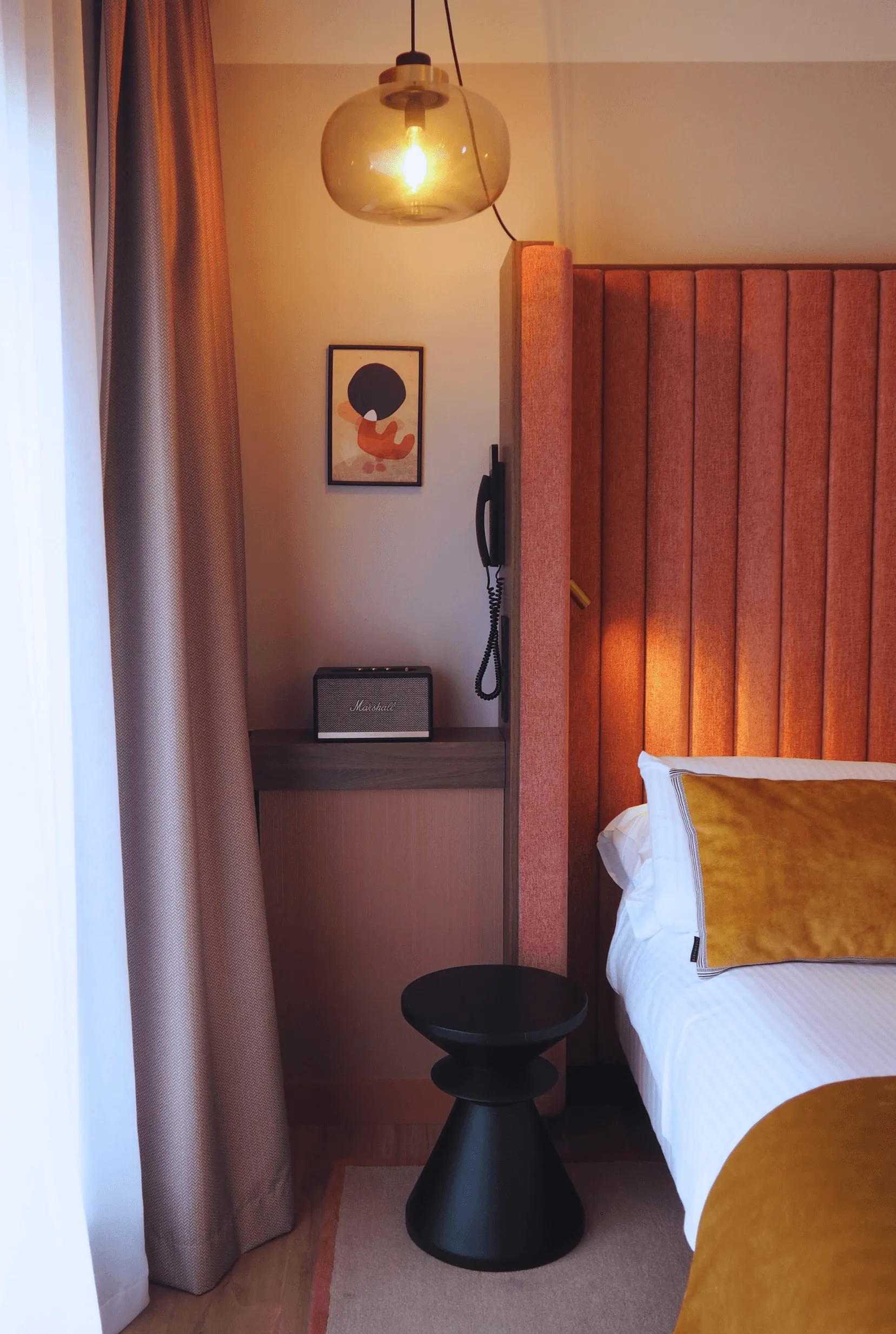 Room with an orange velvet headboard bed, a black bedside table, a radio and a telephone on a shelf, a hanging lamp, and a piece of art on the wall.