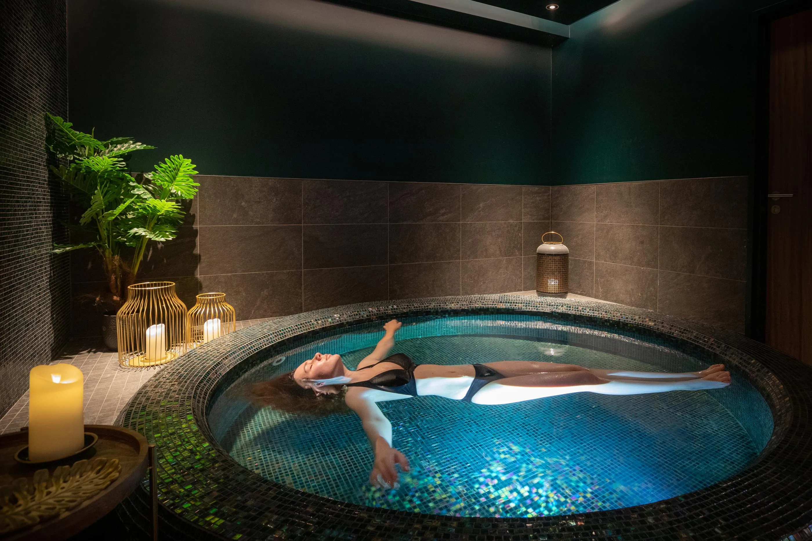 A person lying in a circular hot tub surrounded by candles and green plants.