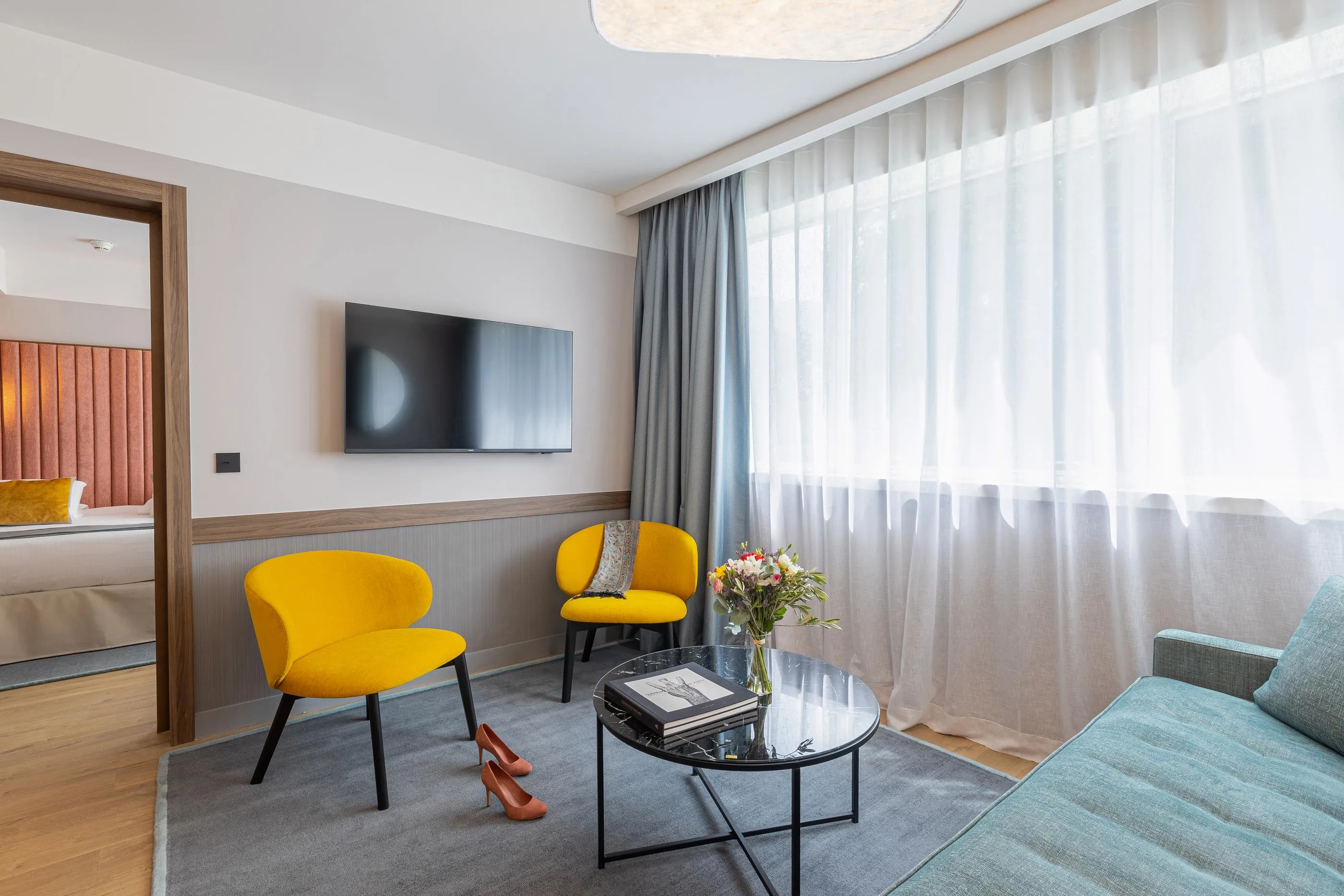 Hotel lounge with two yellow armchairs, a coffee table, magazines, a vase of flowers, and a wall-mounted television.