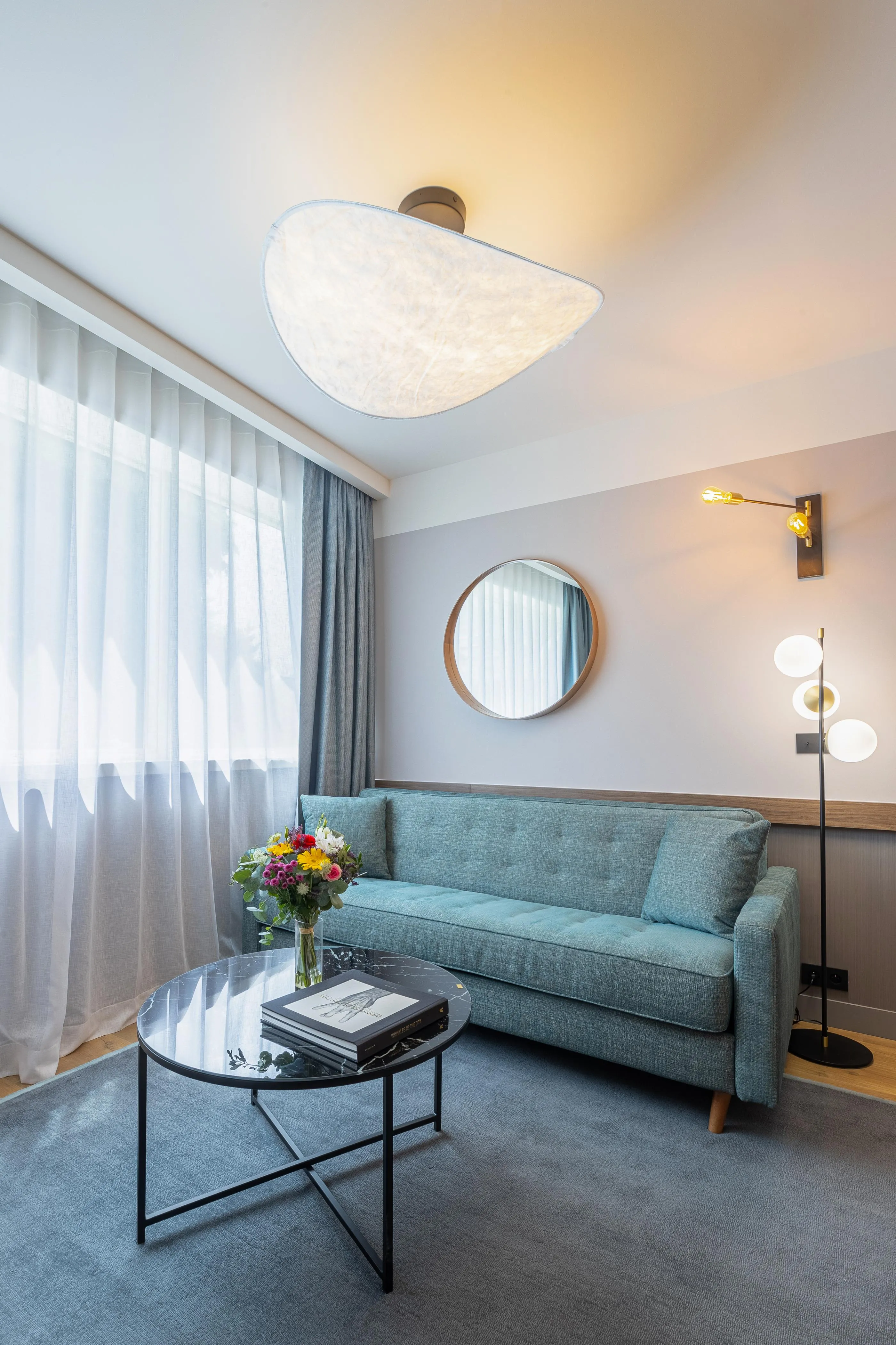 A green sofa, a coffee table with books and flowers, a three-globe lamp, and a round mirror in a modern living room.