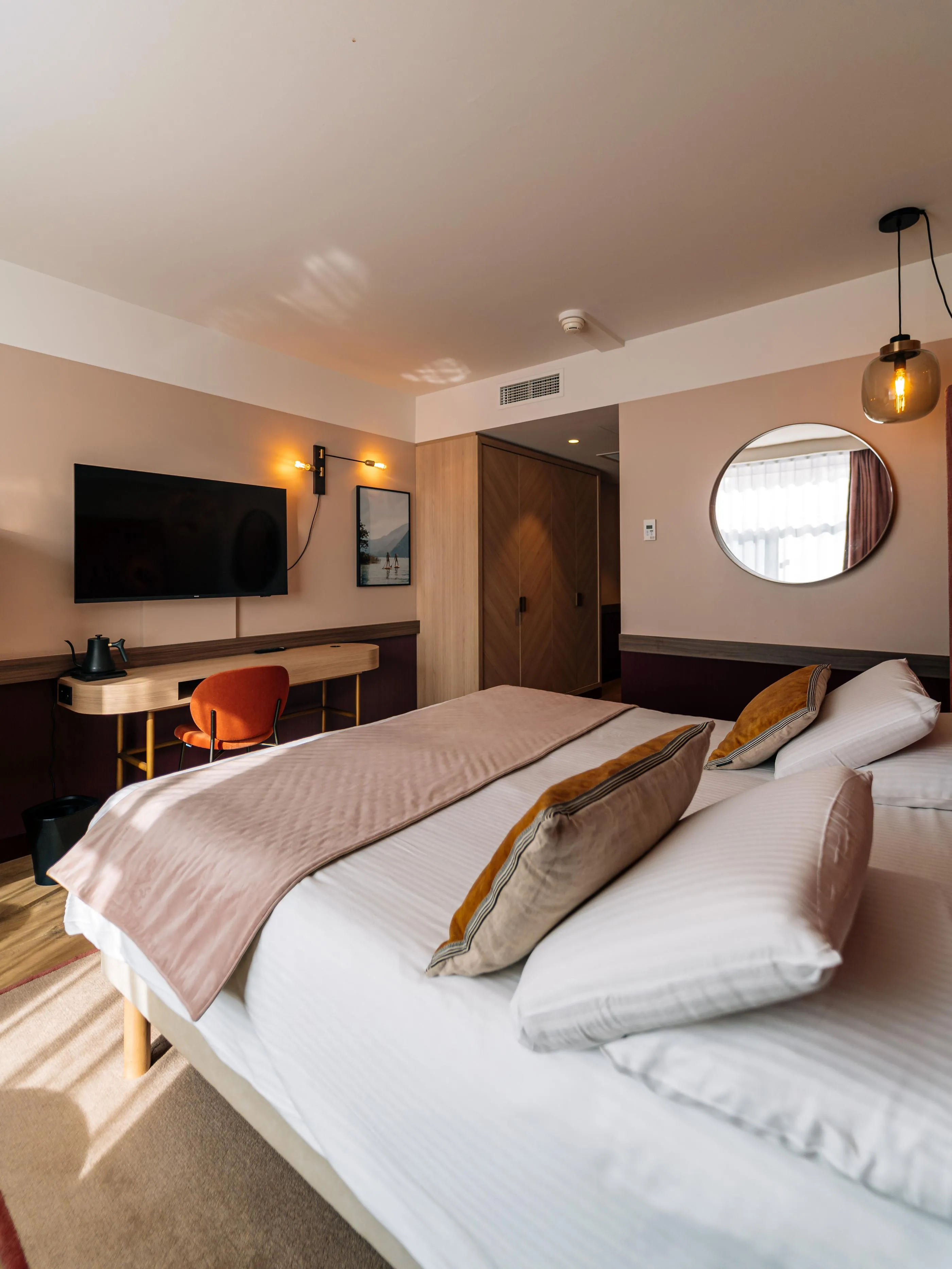 Room with two single beds, a desk, a television, a round mirror, and a hanging lamp.