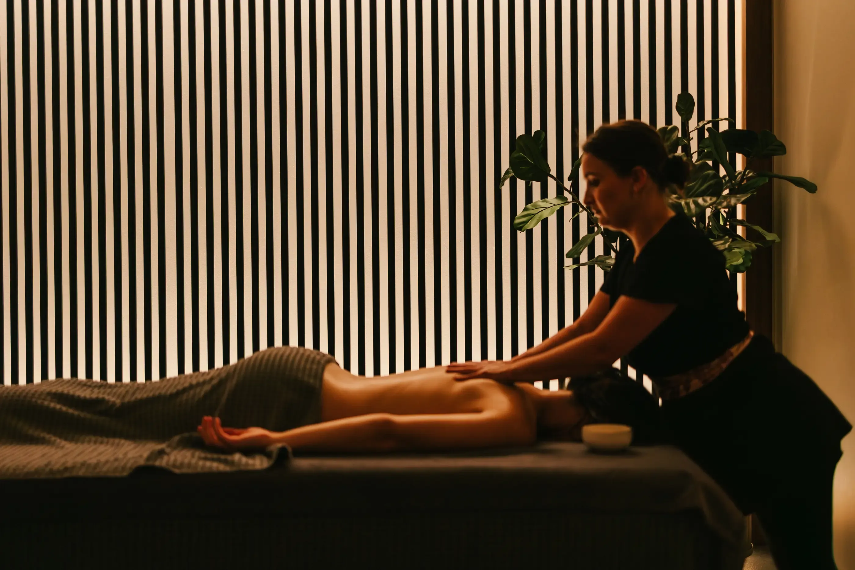 Eine Person erhält eine Massage auf einem Tisch in einem Raum mit einer Pflanze und vertikalen Jalousien.